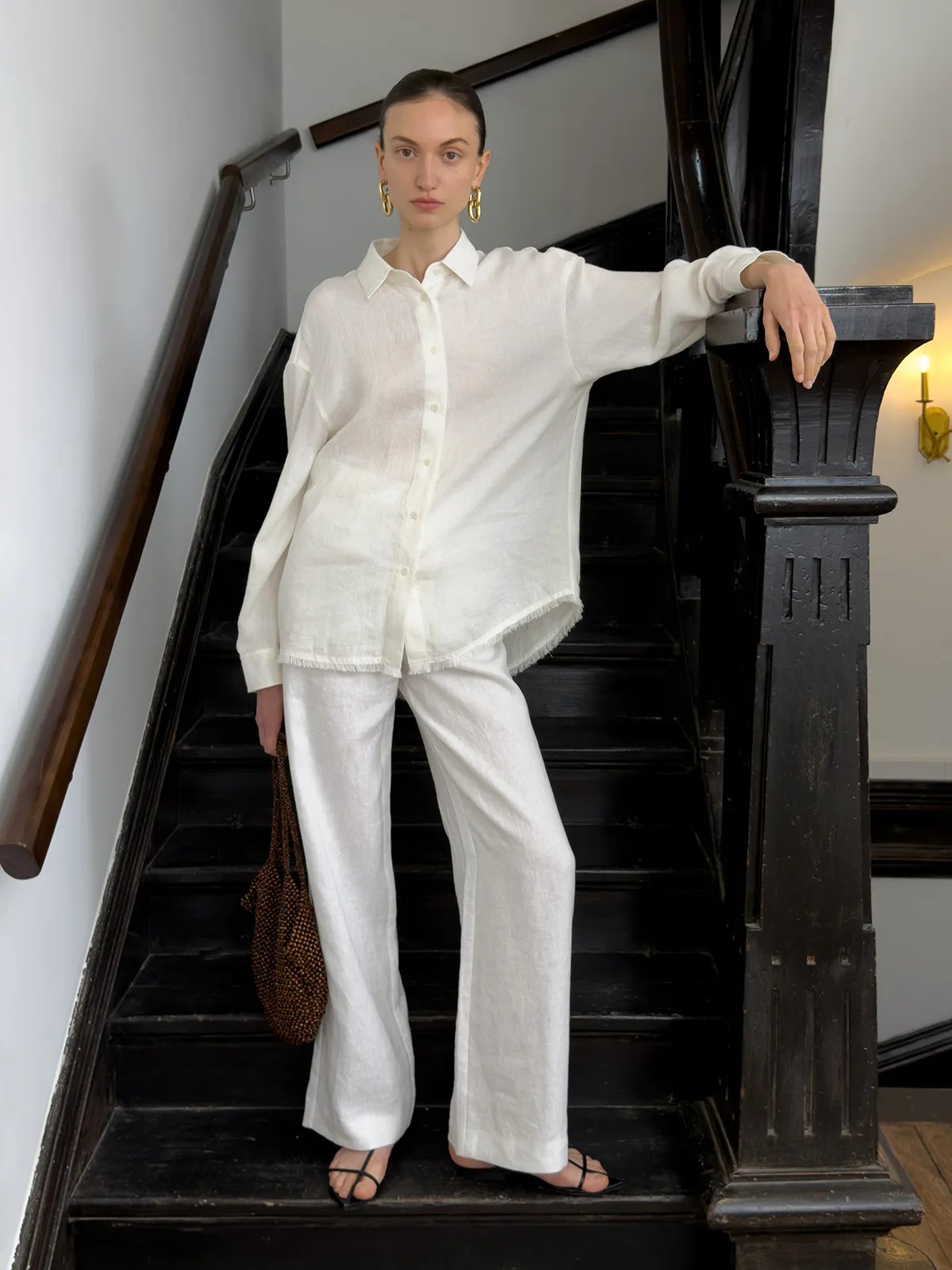 Front-facing model in ivory CLORIS linen trousers and oversized white shirt with frayed hem, holding beaded bag on staircase — editorial minimalism