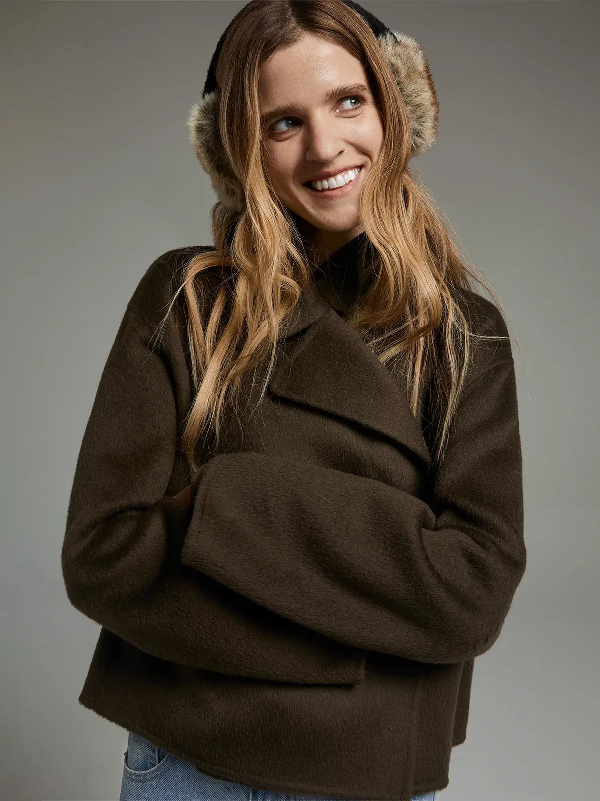 Model wearing a short coat with wide lapels and voluminous sleeves, arms crossed, paired with earmuffs.