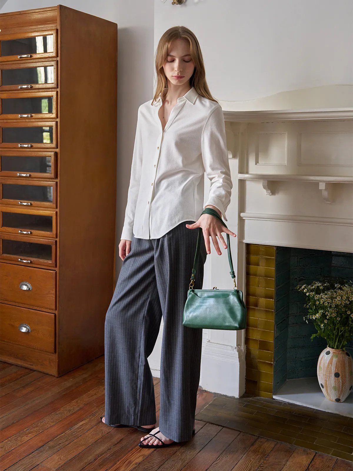 Model holding emerald green leather bag, wearing ivory COLETTE shirt and grey pinstripe trousers in vintage room setting