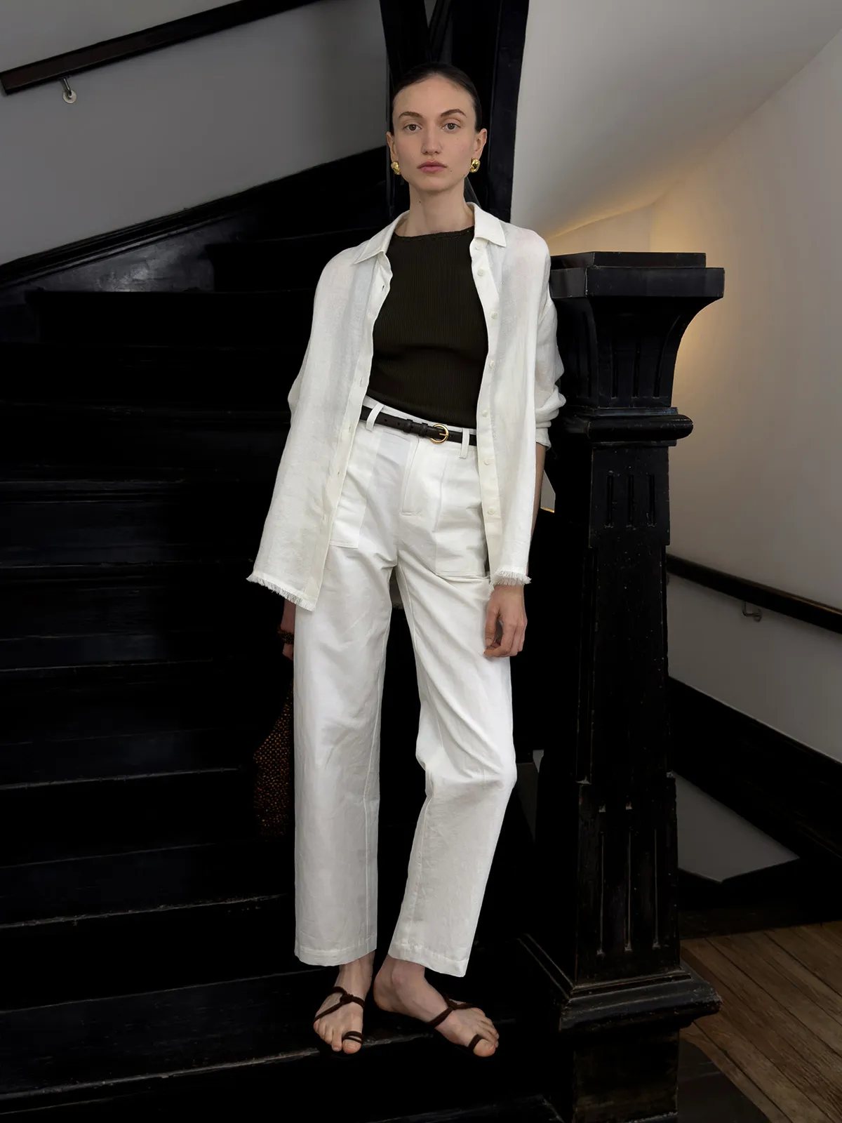 Model standing on black staircase in white REID trousers, layered with ivory shirt and olive top, hands relaxed at sides