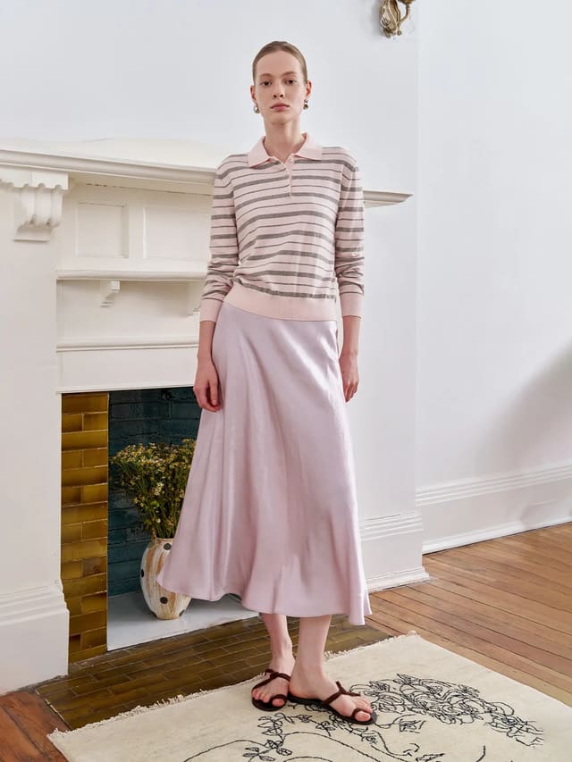 Model wearing SHELL mermaid midi skirt with striped polo shirt, standing by white fireplace in minimalist interior