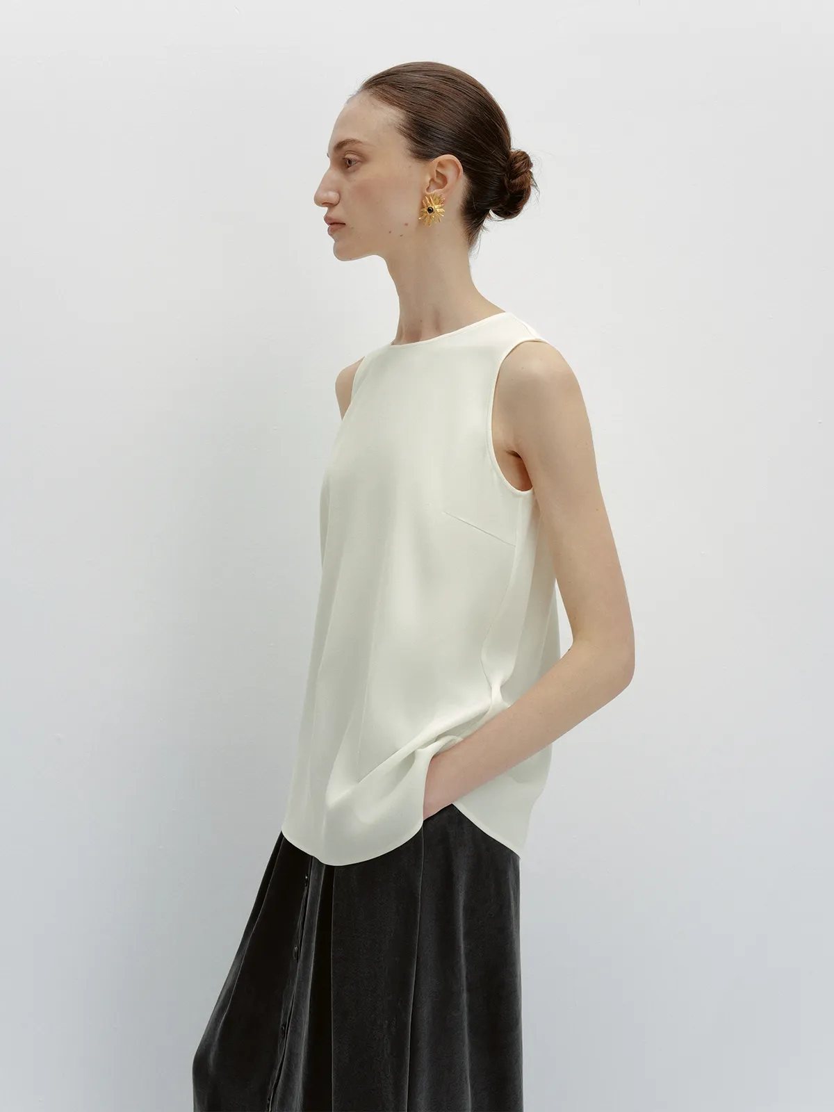 Model in profile wearing ivory MADGE sleeveless blouse with black pleated skirt, gold sunburst earrings, hair in low bun