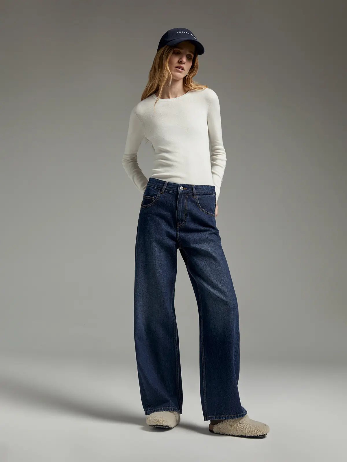 Model wearing a white slim fit long-sleeve knit top with crew neck, dark jeans, and a cap.