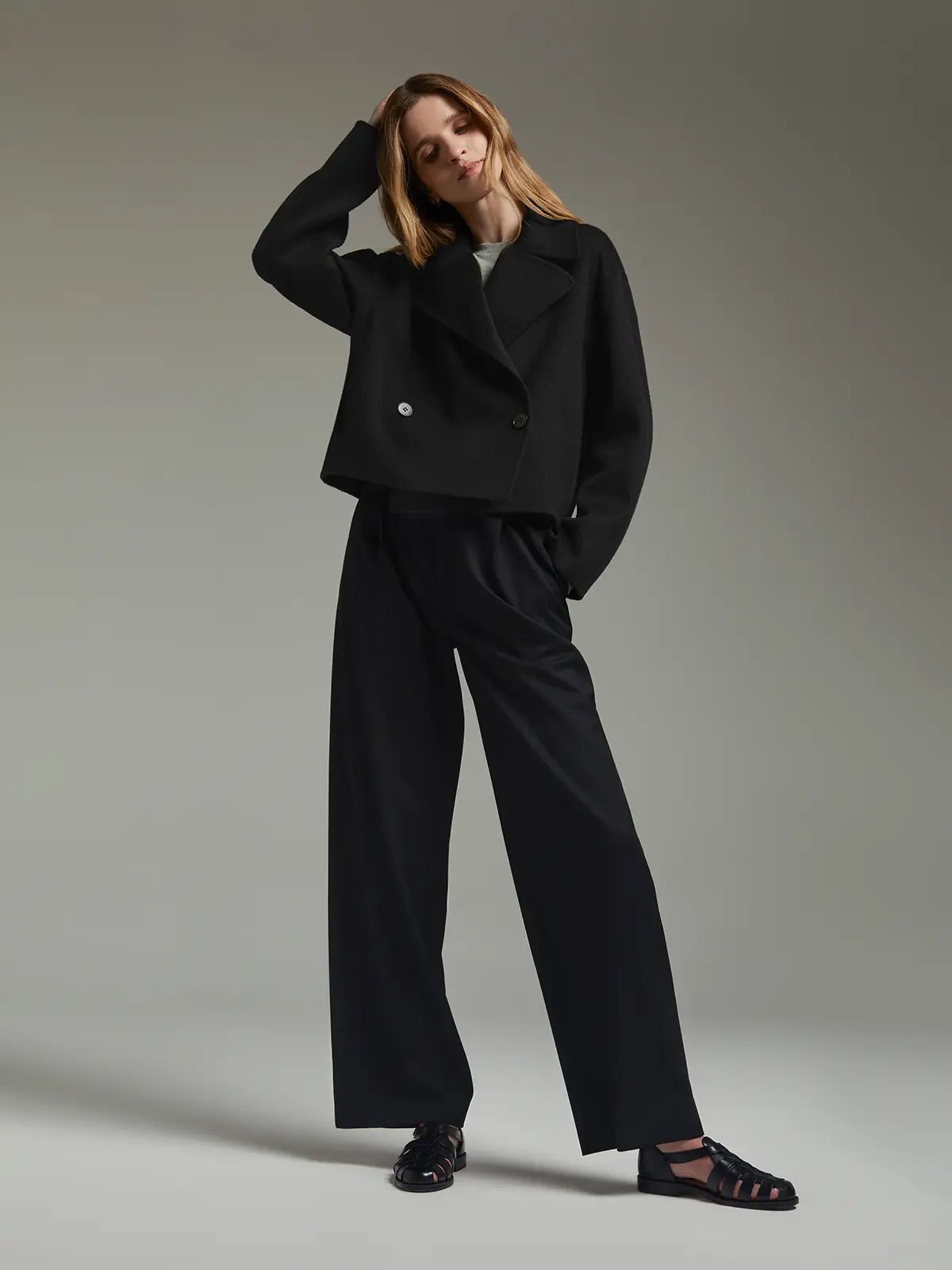 Model wearing a short double-breasted coat with wide lapels and flared sleeves, one hand in hair, paired with trousers.