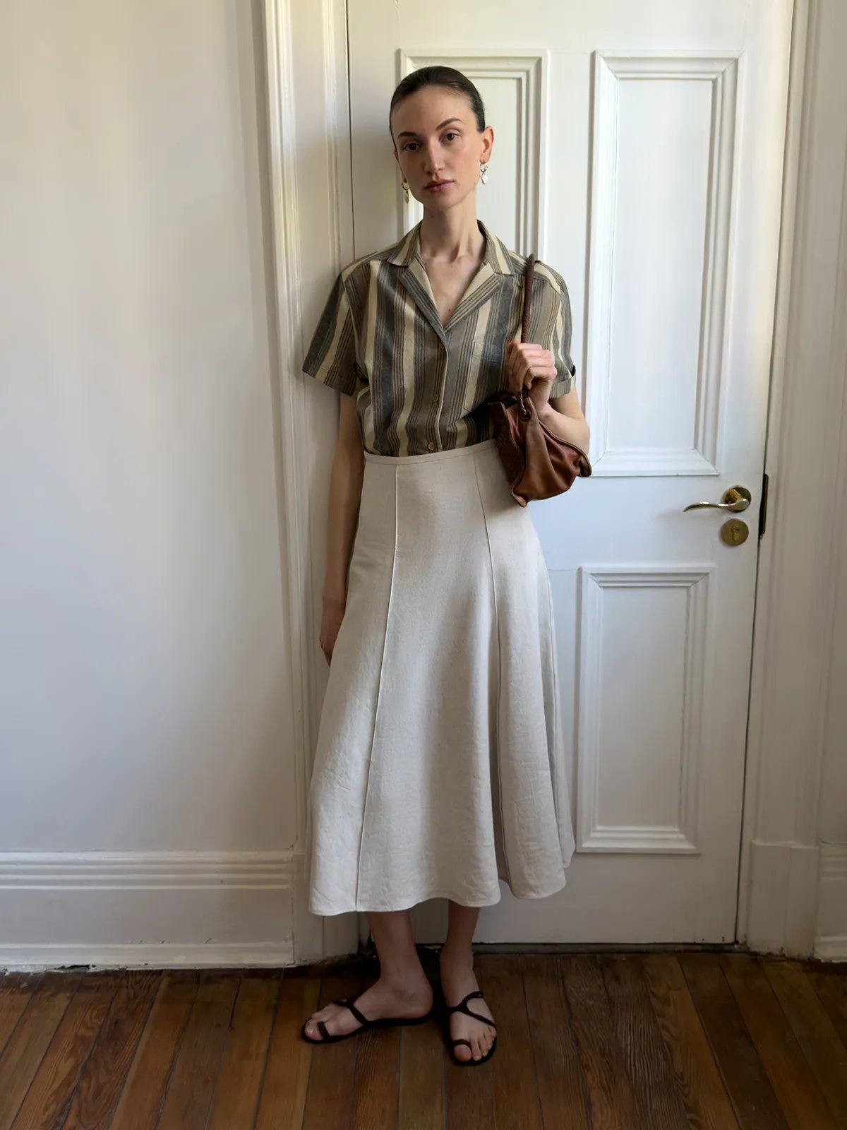 Model leaning against white door in NATASHA skirt, striped shirt, and brown leather bag