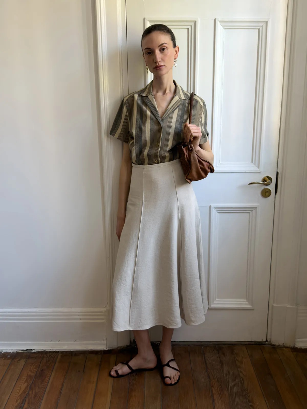Model leaning against white door in NATASHA skirt, striped shirt, and brown leather bag