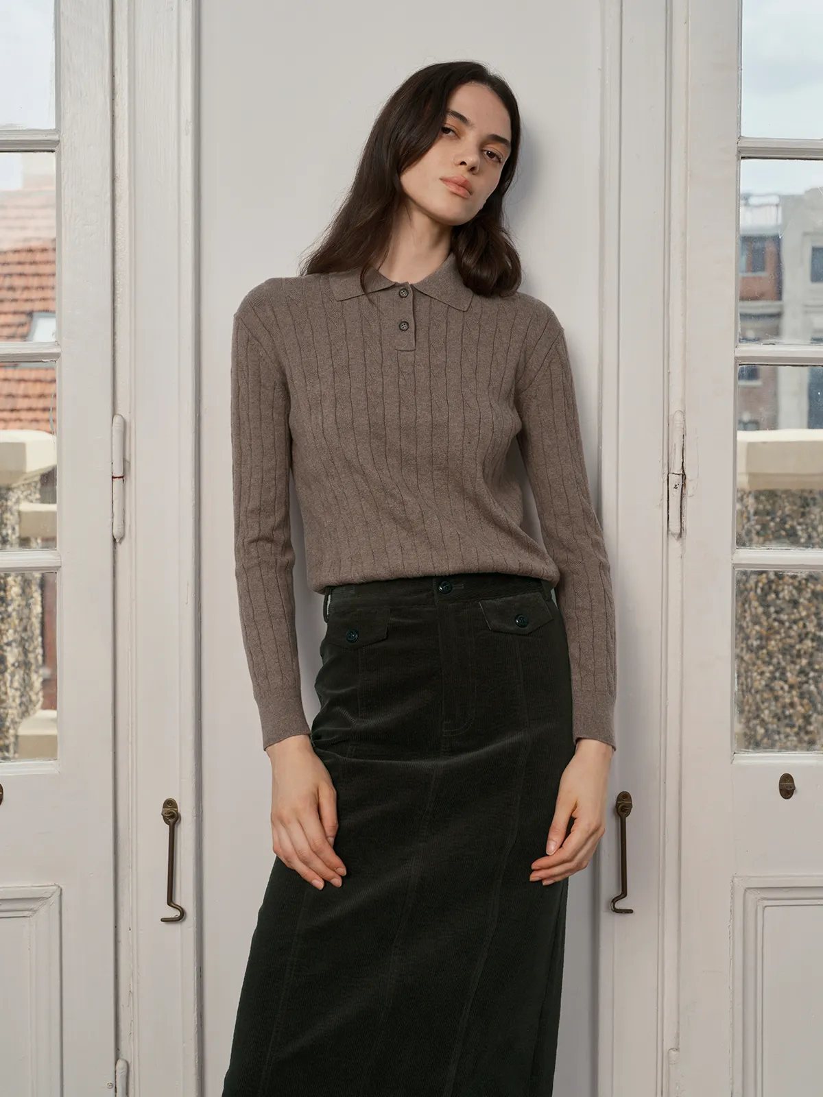 Model in taupe EMERSON Cable Knit Polo Jumper with dark green corduroy midi skirt, standing by white French doors—minimalist autumn outfit