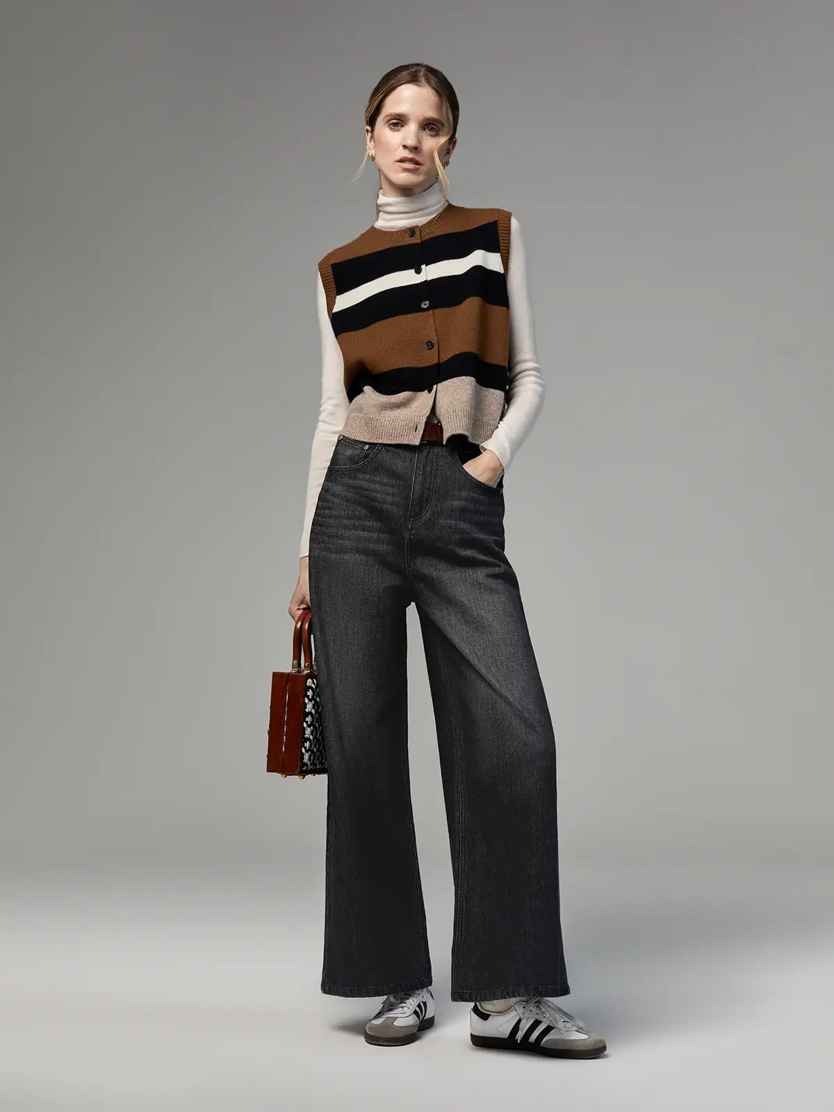 Model wearing Dark Grey wide-leg jeans with a striped vest, turtleneck, and sneakers, holding a patterned handbag.