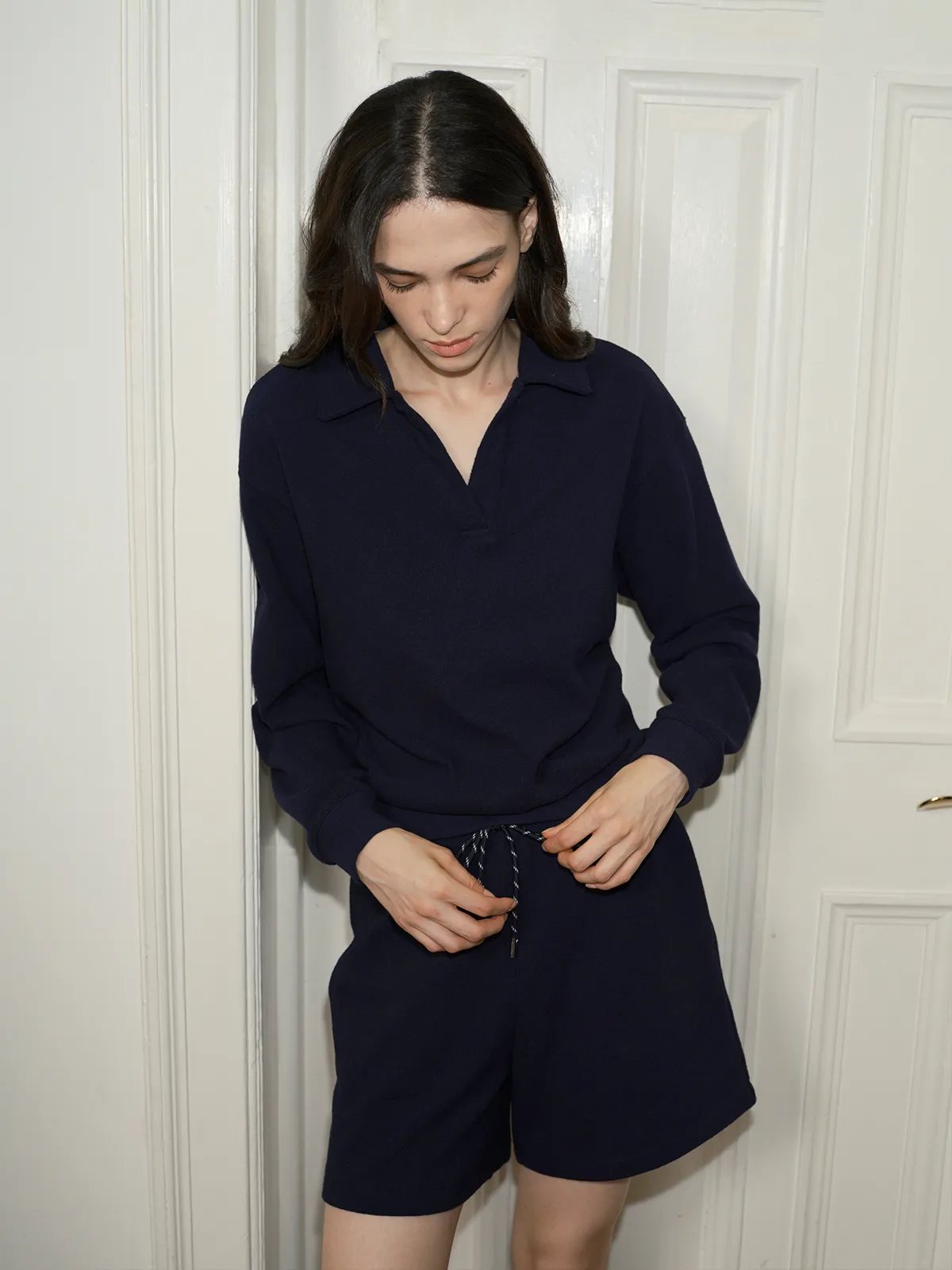 Model adjusting drawstring on navy CASPER sweatshirt and shorts set against white door