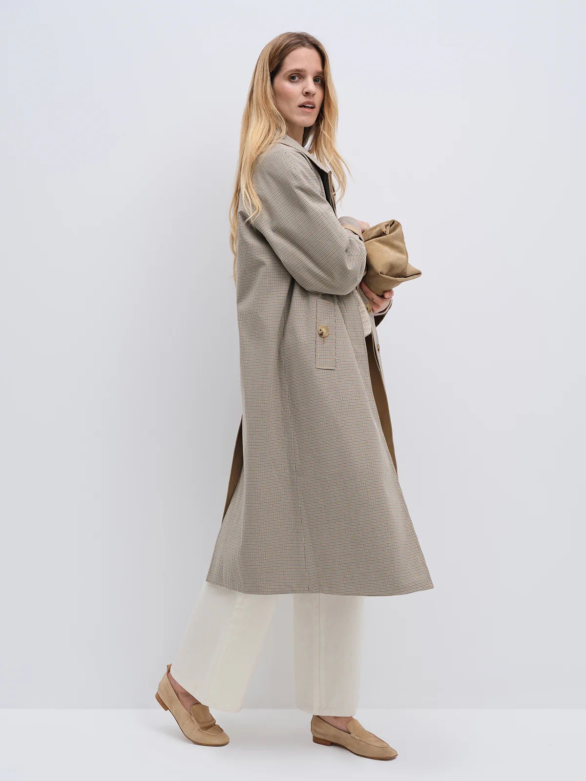 Side view of a model in a Khaki reversible check trench coat with hardware, worn over a knit top and trousers.