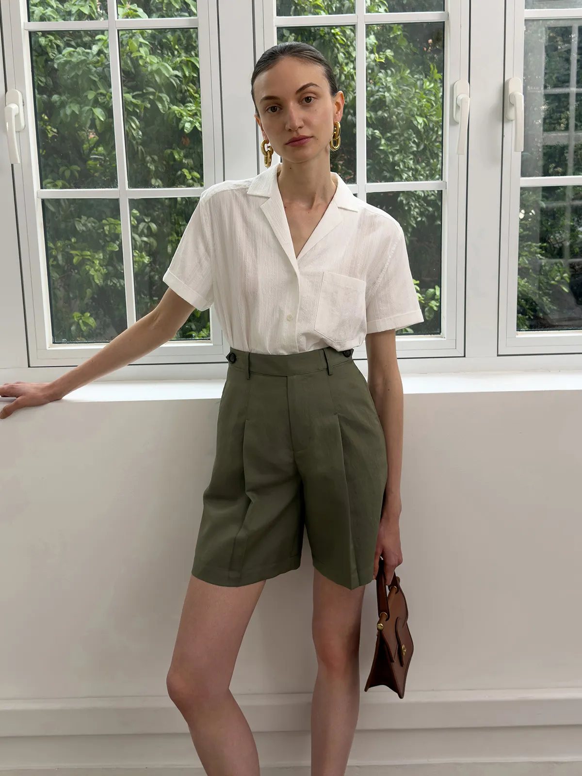 Model wearing ISADORA white shirt with olive green pleated high-waisted shorts by window
