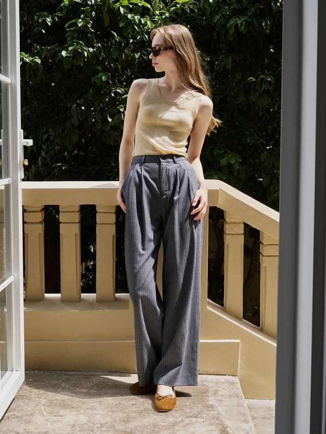 Model wearing oat MEADOW Textured Knit Tank Top with grey pinstripe wide-leg trousers on sunlit balcony, embodying minimalist summer elegance