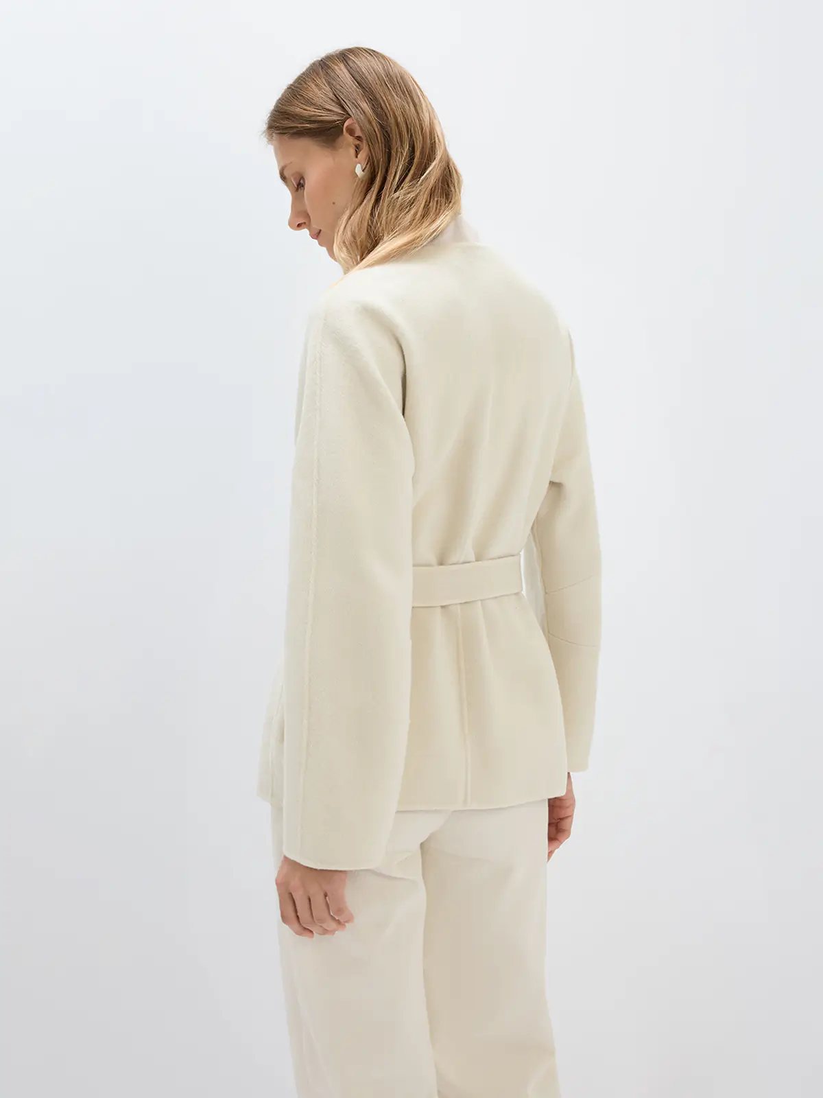 Side view of a model in an off-white belted coat with long sleeves, wrap closure, and waist tie.
