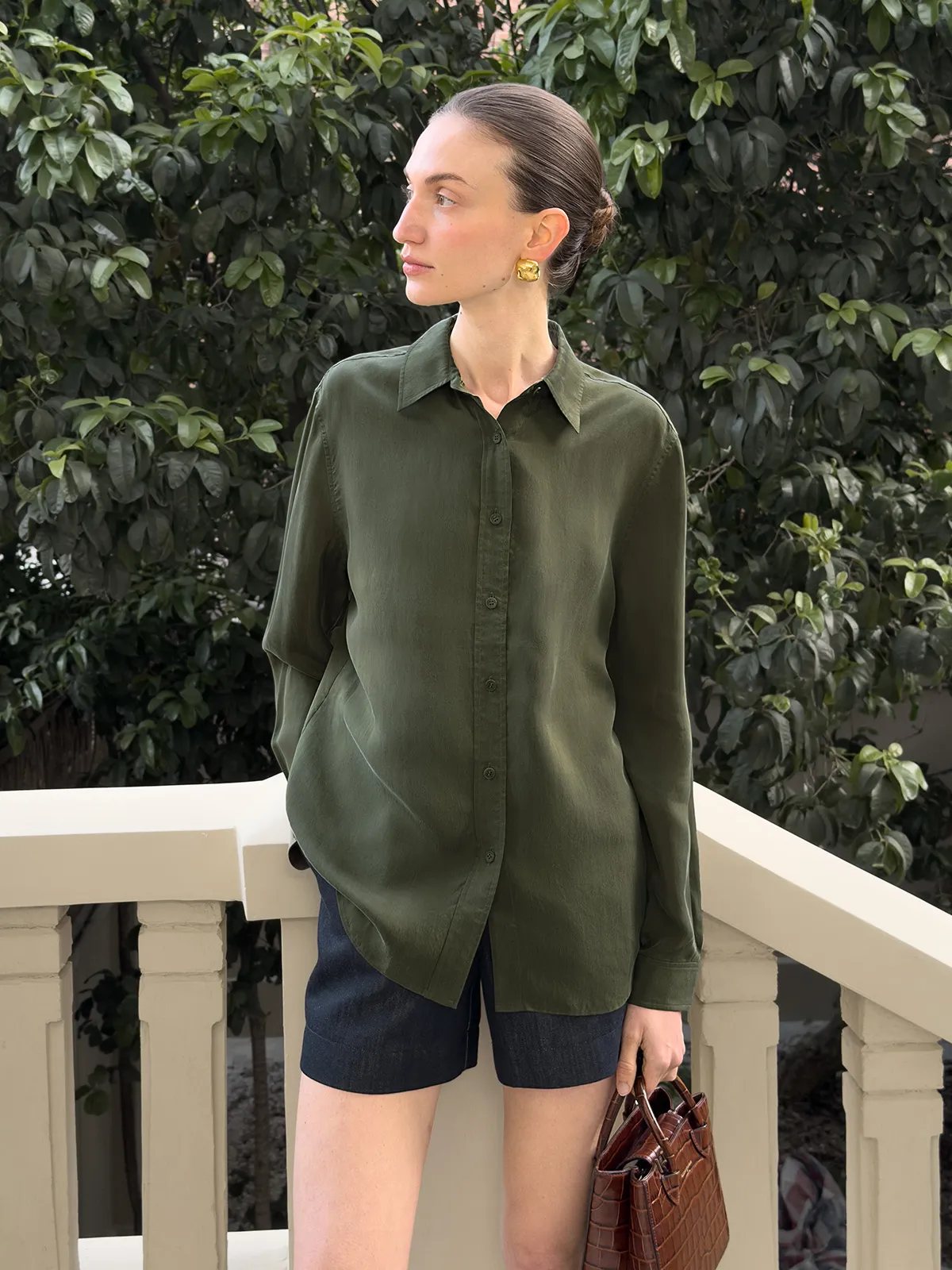 Model wearing GREER olive cupro shirt with navy denim shorts and croc-embossed bag on balcony