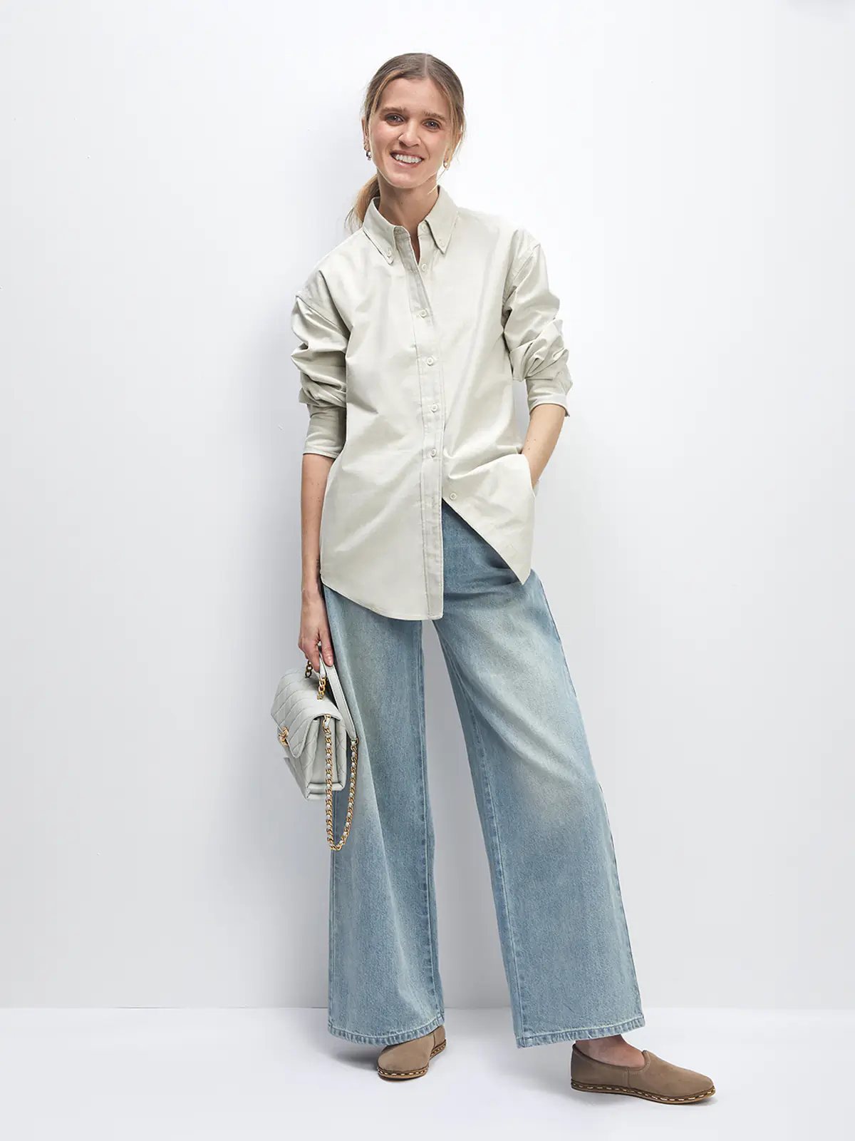 Side profile of a model wearing the loose Oxford shirt in Slate Gray with wide-leg jeans and holding a quilted light gray handbag.