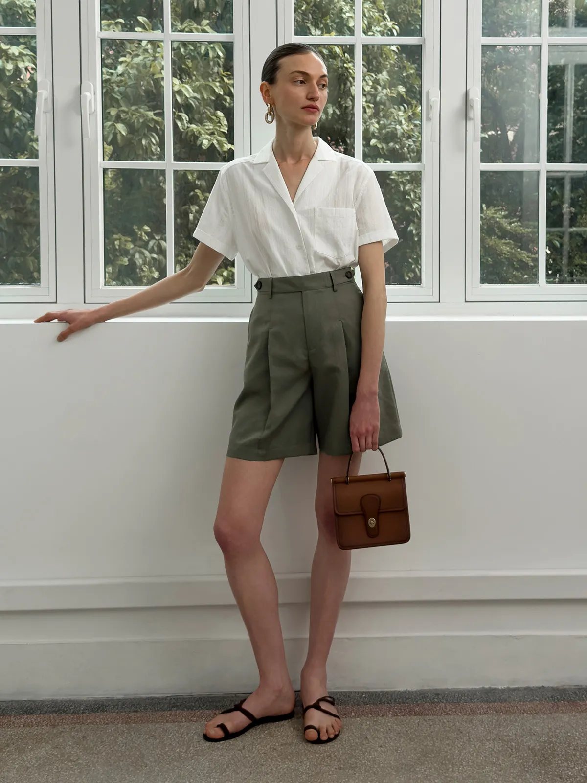 Model wearing olive green ADELE Bermuda shorts with white short-sleeve shirt and brown leather bag by window