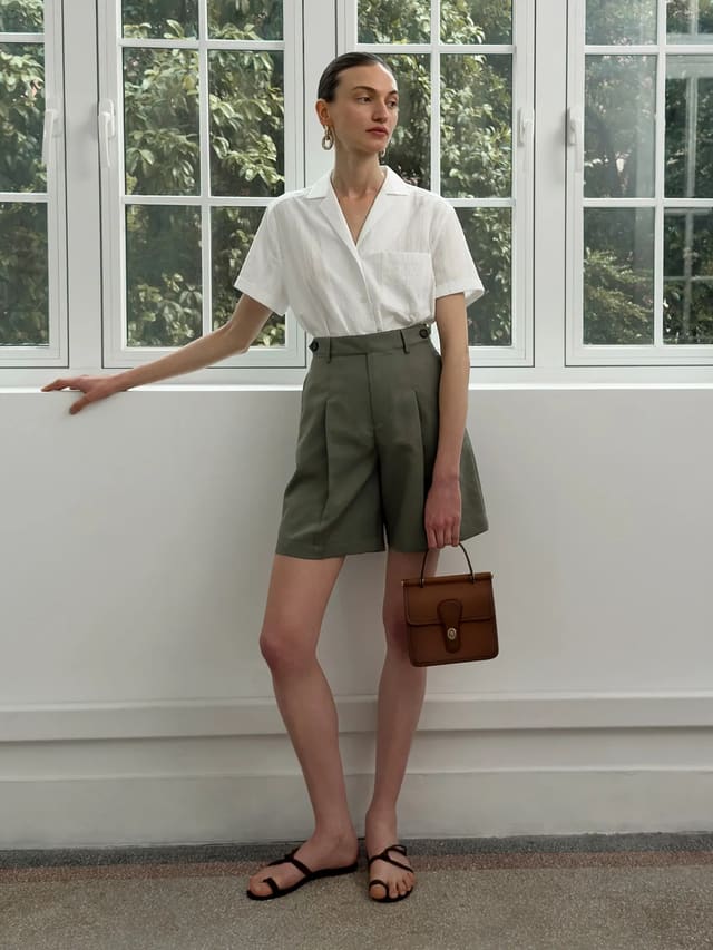 Model wearing olive green ADELE Bermuda shorts with white short-sleeve shirt and brown leather bag by window