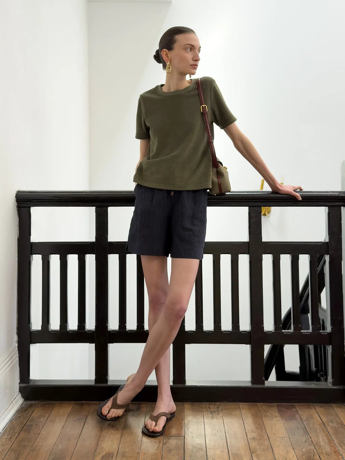 Model in olive LANE Terry tee with navy corduroy shorts and brown leather crossbody bag on indoor staircase