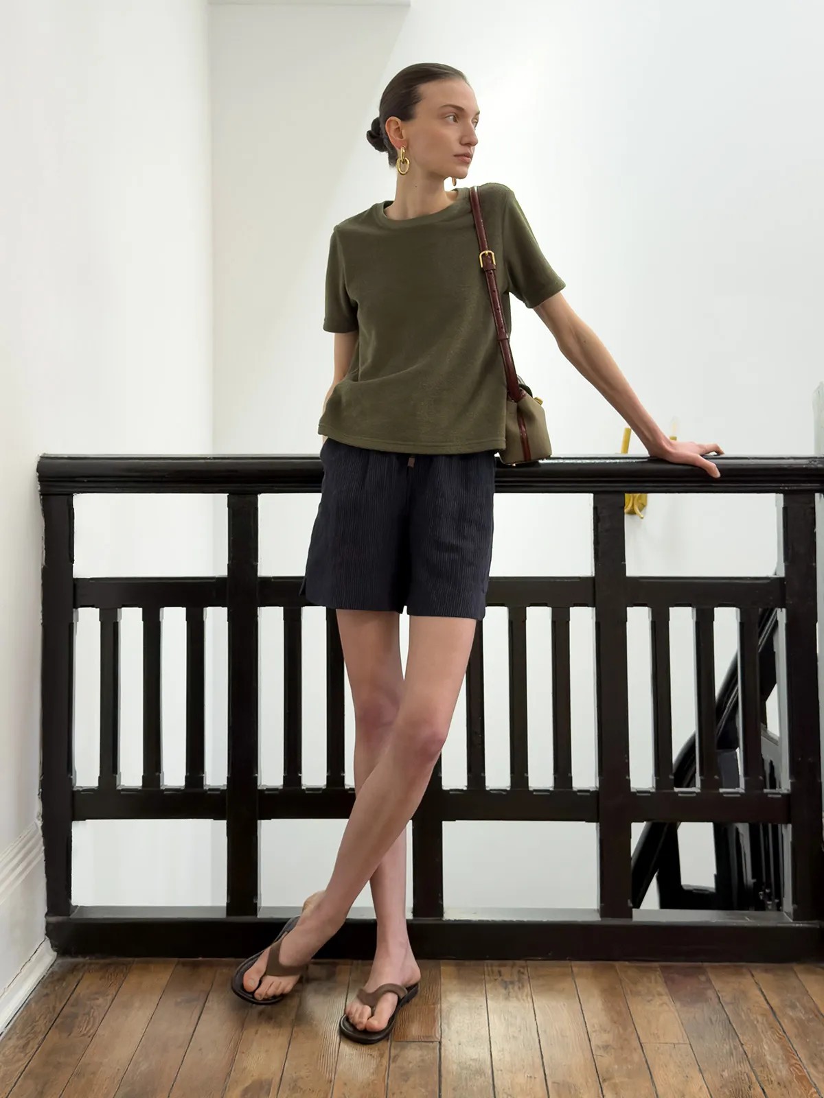 Model in olive LANE Terry tee with navy corduroy shorts and brown leather crossbody bag on indoor staircase