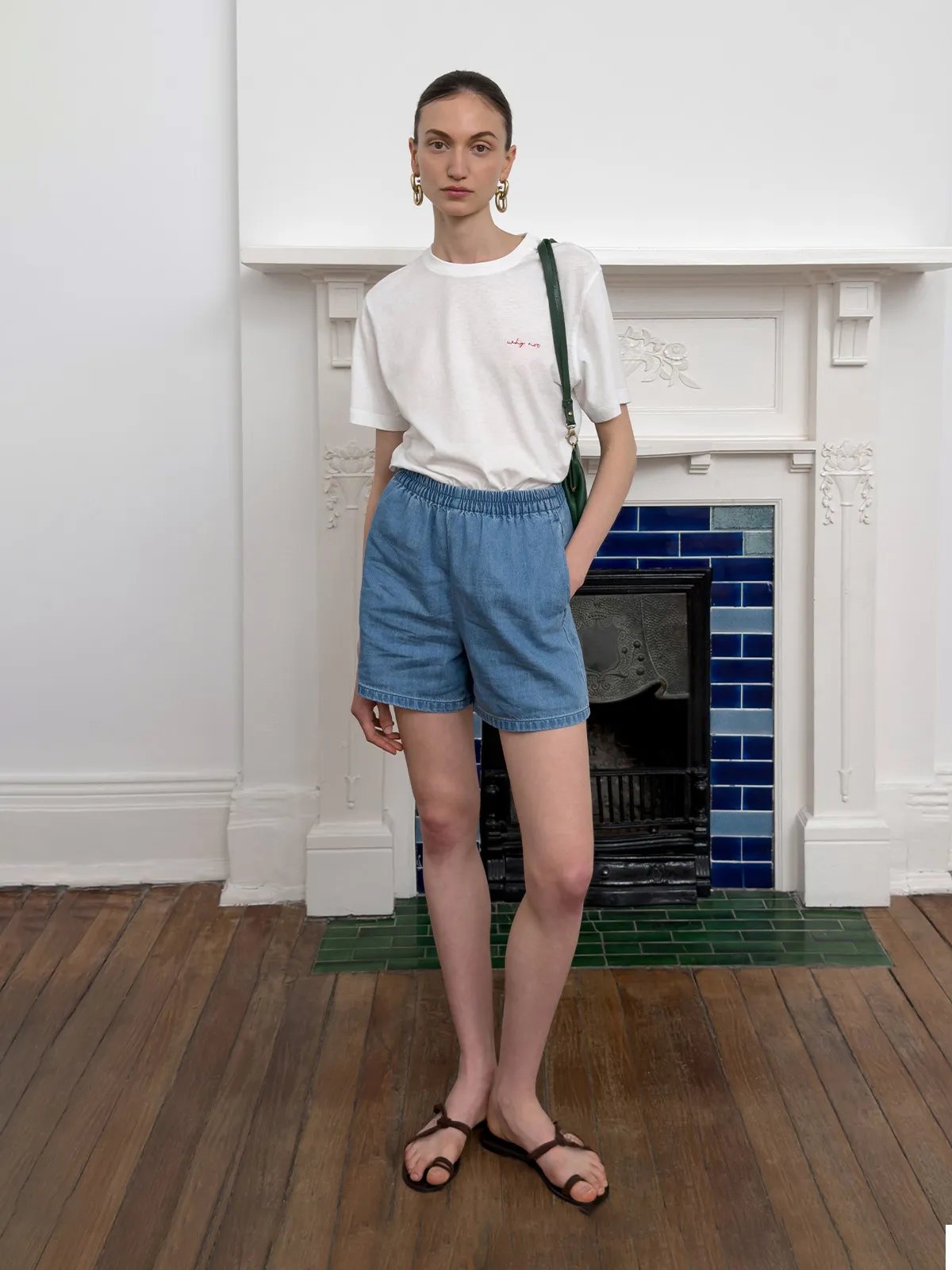 Model wearing WAYLON denim shorts with white embroidered tee, green leather bag, and brown strappy sandals in front of ornate white fireplace