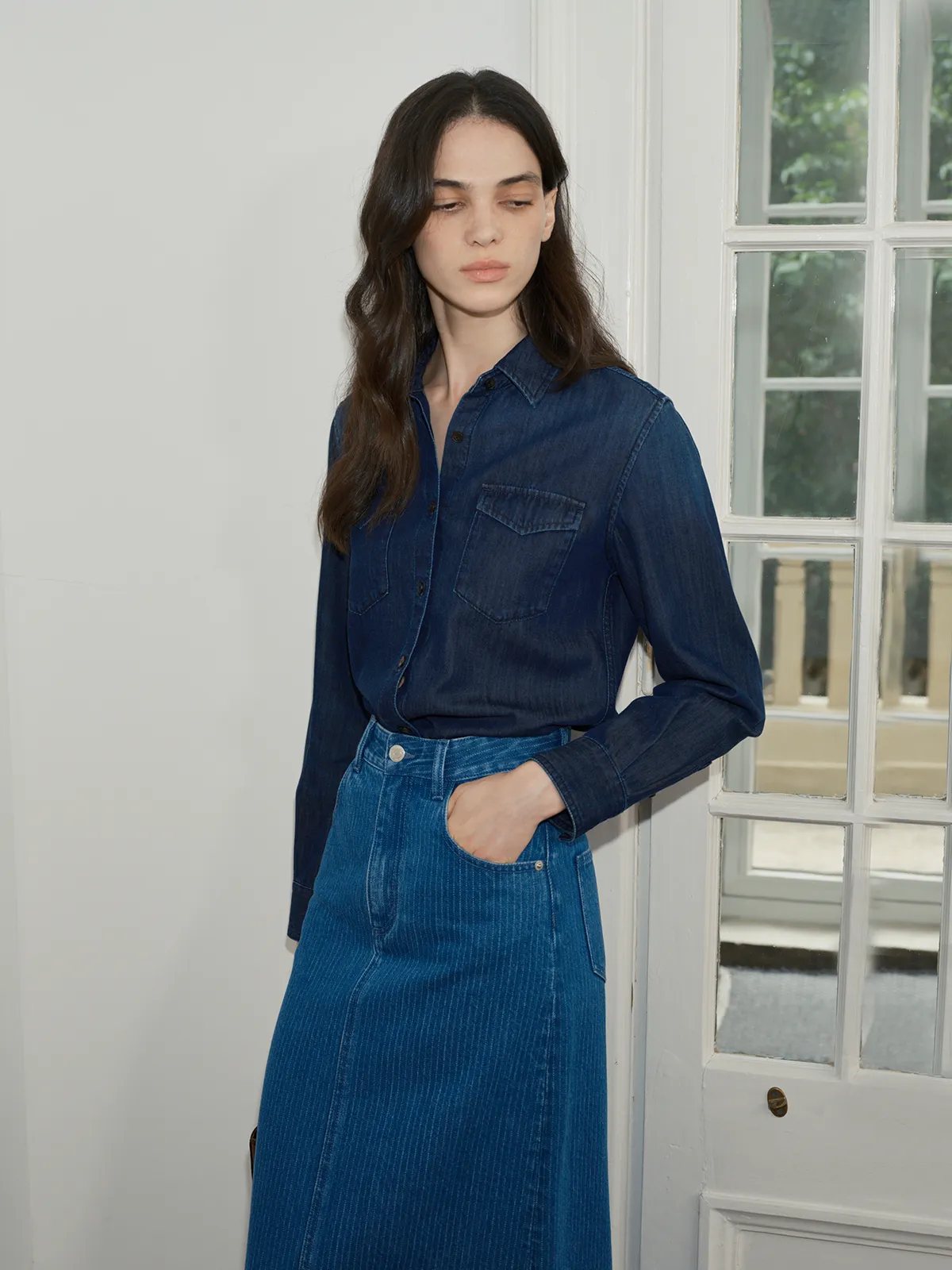 Side profile of model in BRYCE denim maxi skirt with matching denim shirt, hand in pocket, near French window