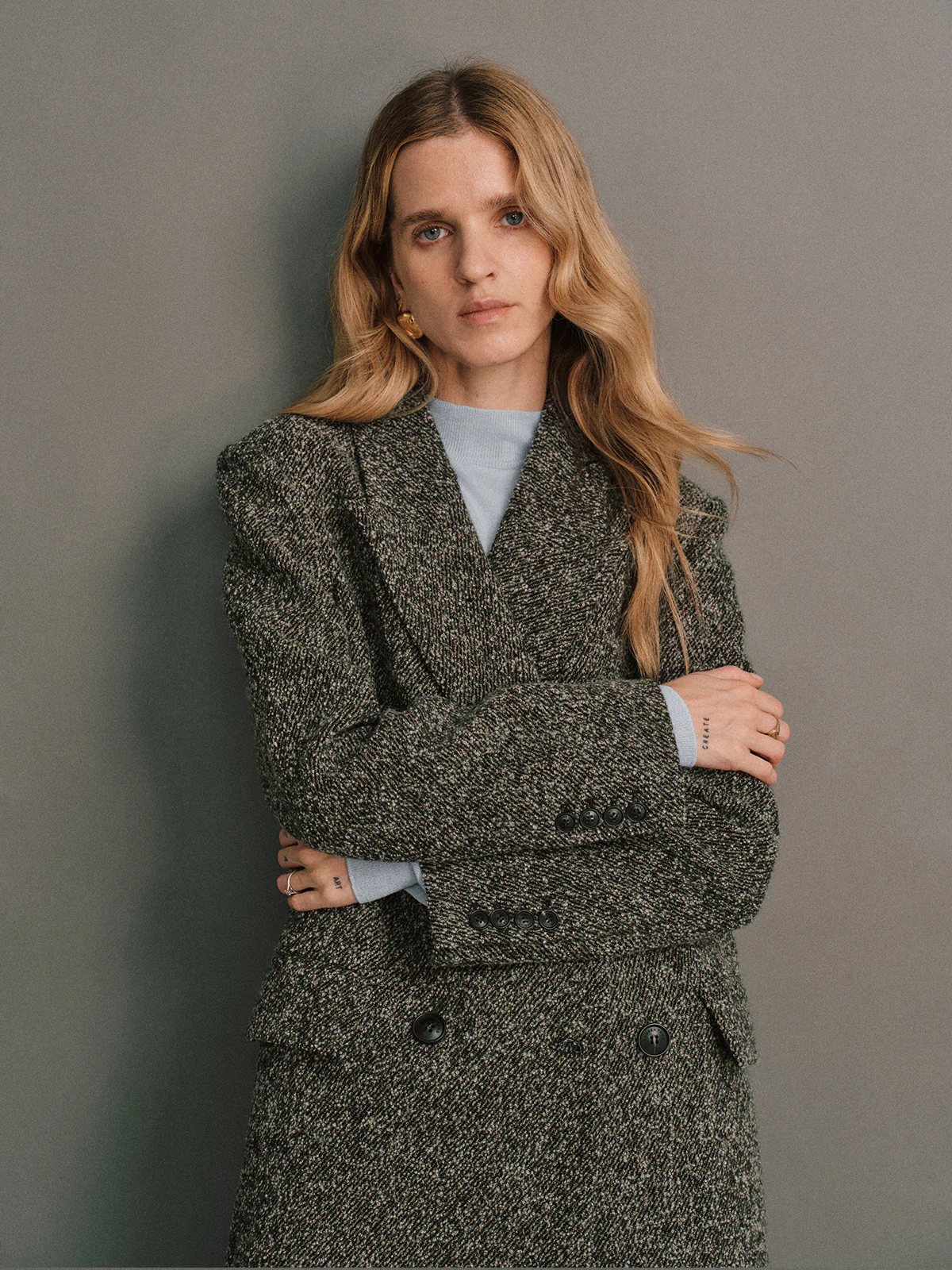 Front view of a model in a Gray wool bouclé tweed tailored long coat, double-breasted, worn over a turtleneck and gray skirt, hand resting on hip.