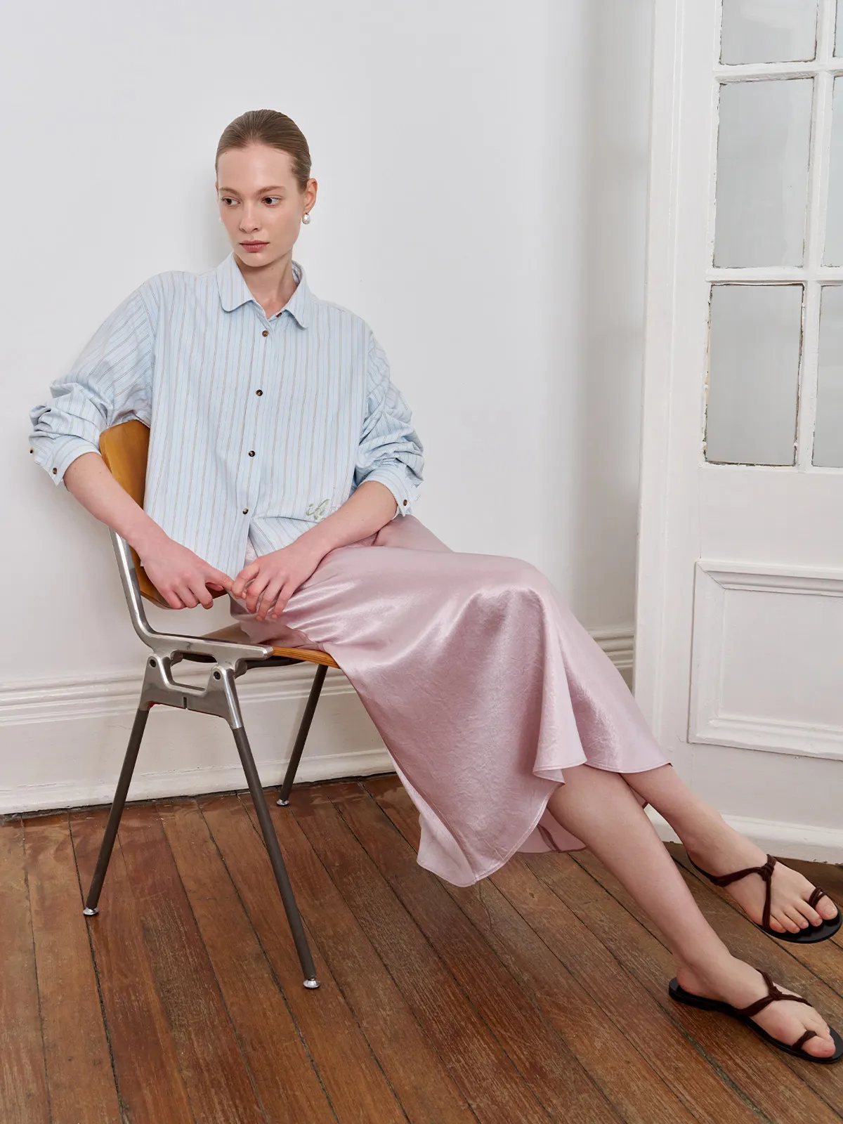Model seated on modern chair wearing SHELL skirt with light blue striped shirt and brown sandals