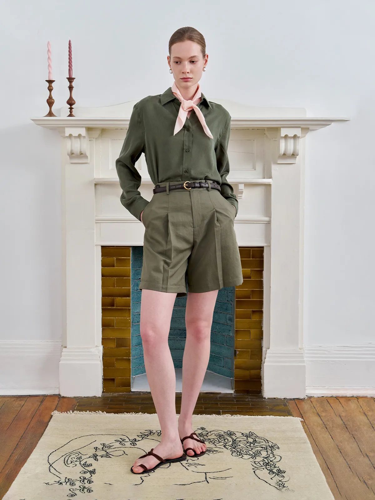 Model in GREER shirt with pink silk scarf, pleated olive shorts, and belt in vintage fireplace setting