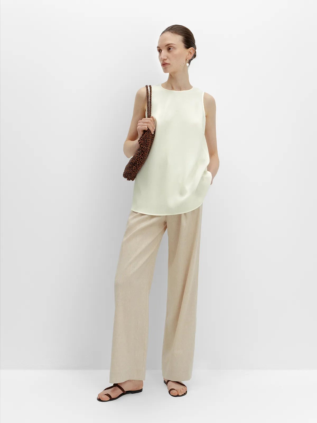 Full-length side pose: model in ivory MADGE blouse, beige wide-leg pants, brown leather sandals, holding beaded clutch