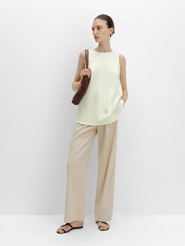 Full-length side pose: model in ivory MADGE blouse, beige wide-leg pants, brown leather sandals, holding beaded clutch