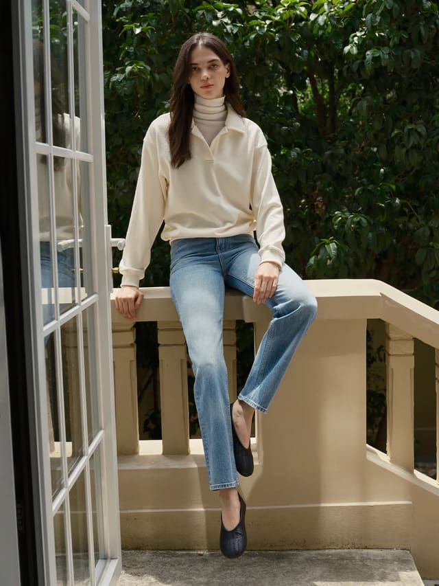 Model wearing ivory CASPER relaxed polo sweatshirt with turtleneck and wide-leg jeans on balcony