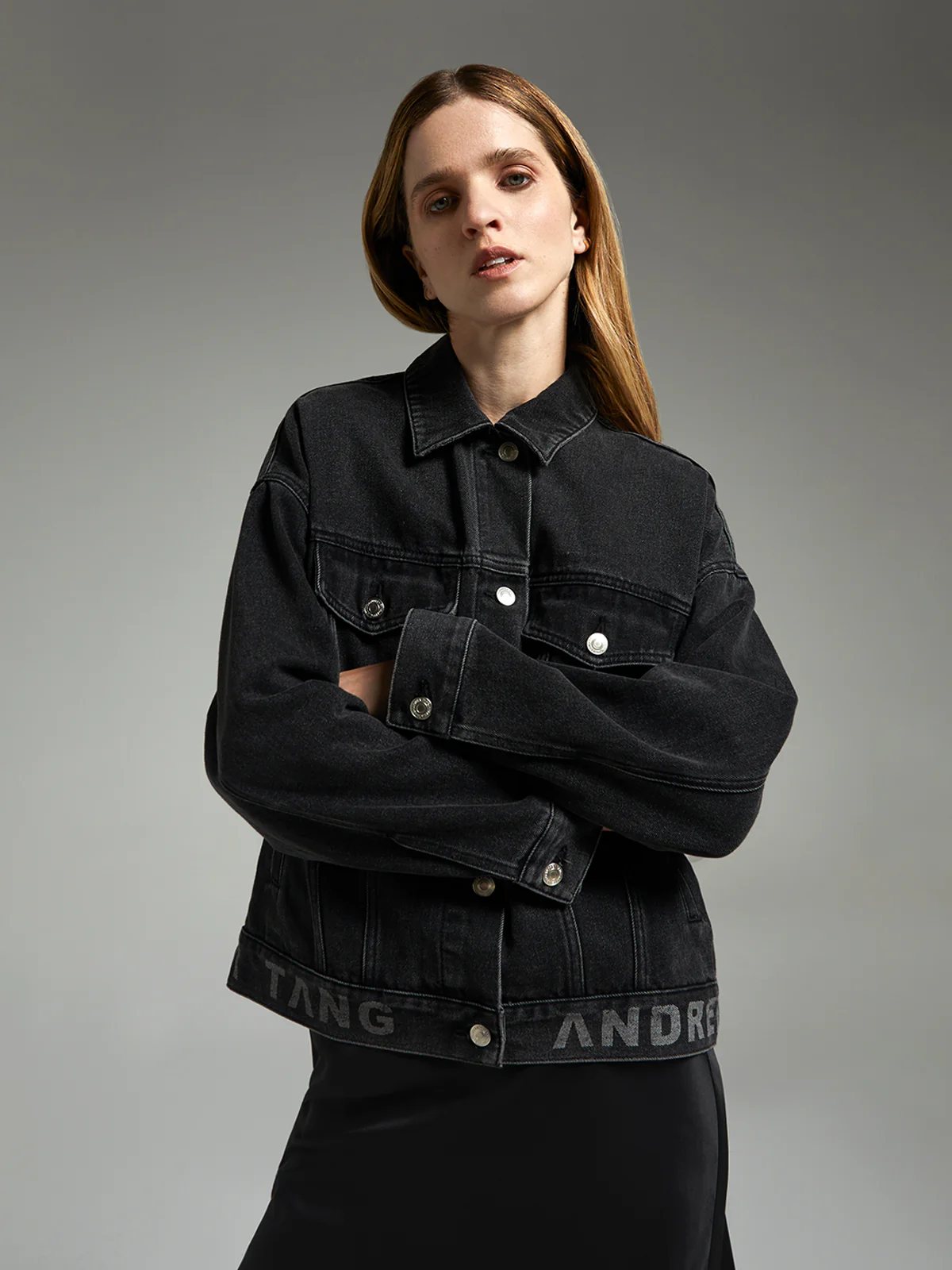 Close-up of andreatang washed-black denim jacket showing chest pockets, metal buttons and tonal logo jacquard at hem