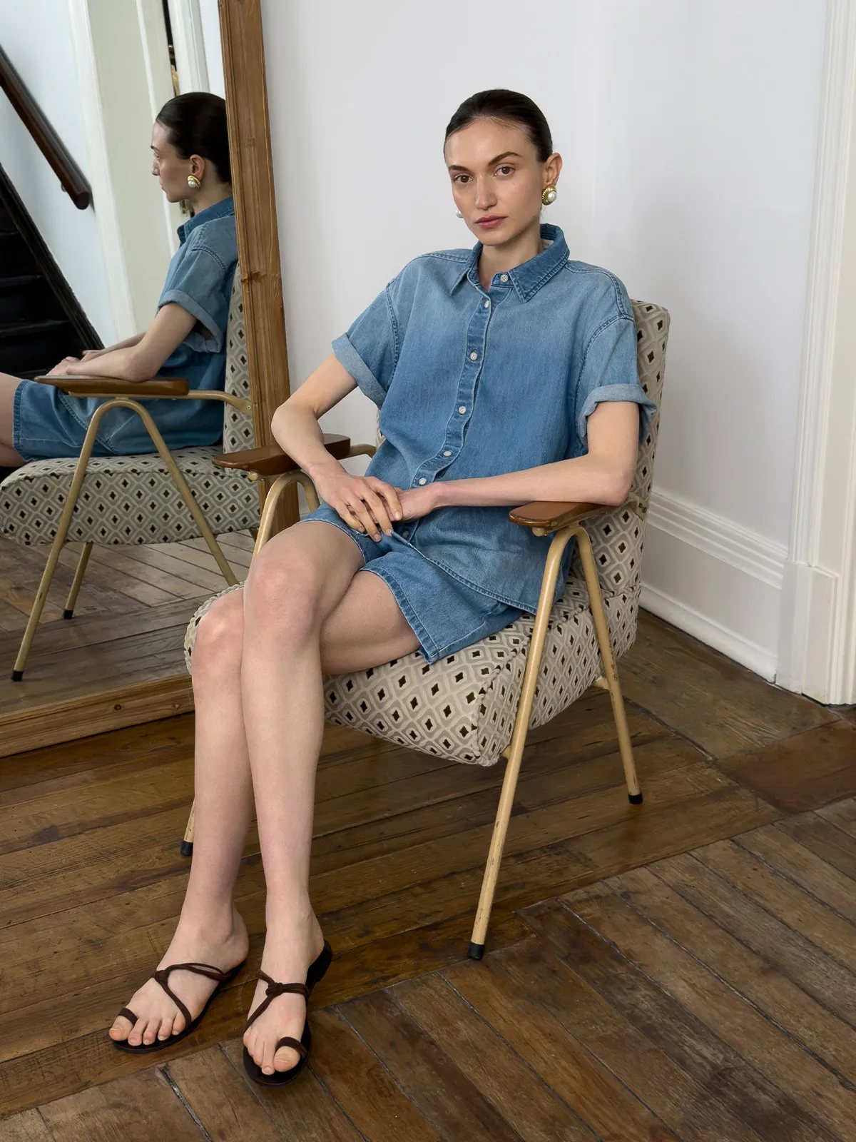 Model seated in mid-century chair wearing denim shirt-dress with mirror reflection