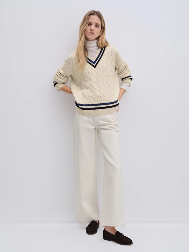 Off-white cable knit V-neck tennis jumper with contrasting trim, worn over a turtleneck with hands in pockets.