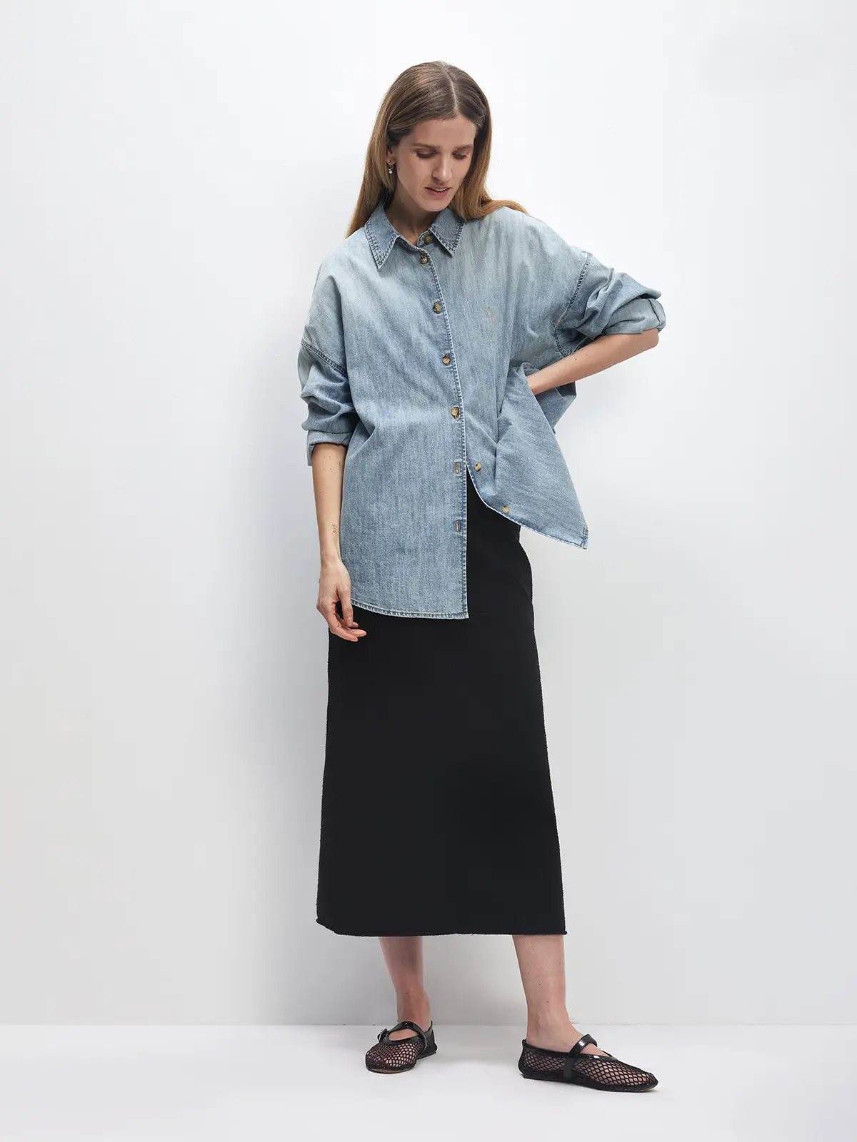 Model wears a light blue oversized denim shirt with rolled sleeves over a midi skirt, hands on hips.