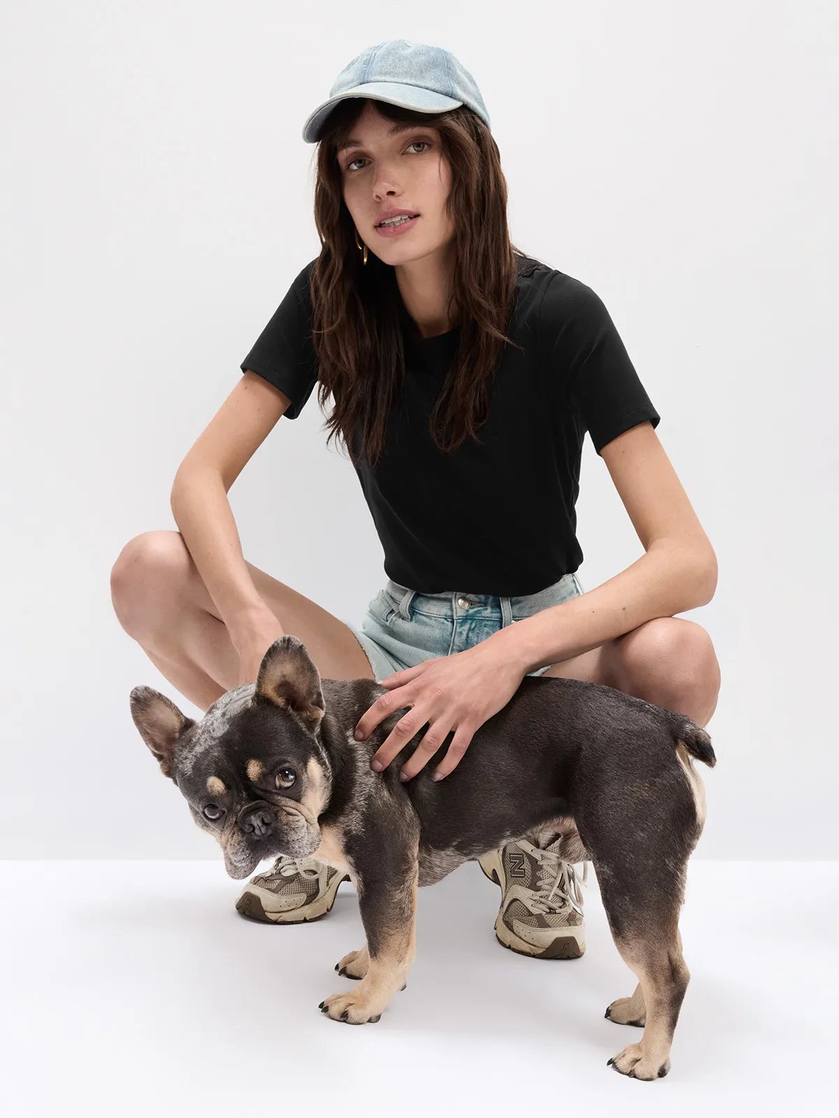 Model crouching with French bulldog, wearing black SOSA t-shirt, denim shorts, and denim cap