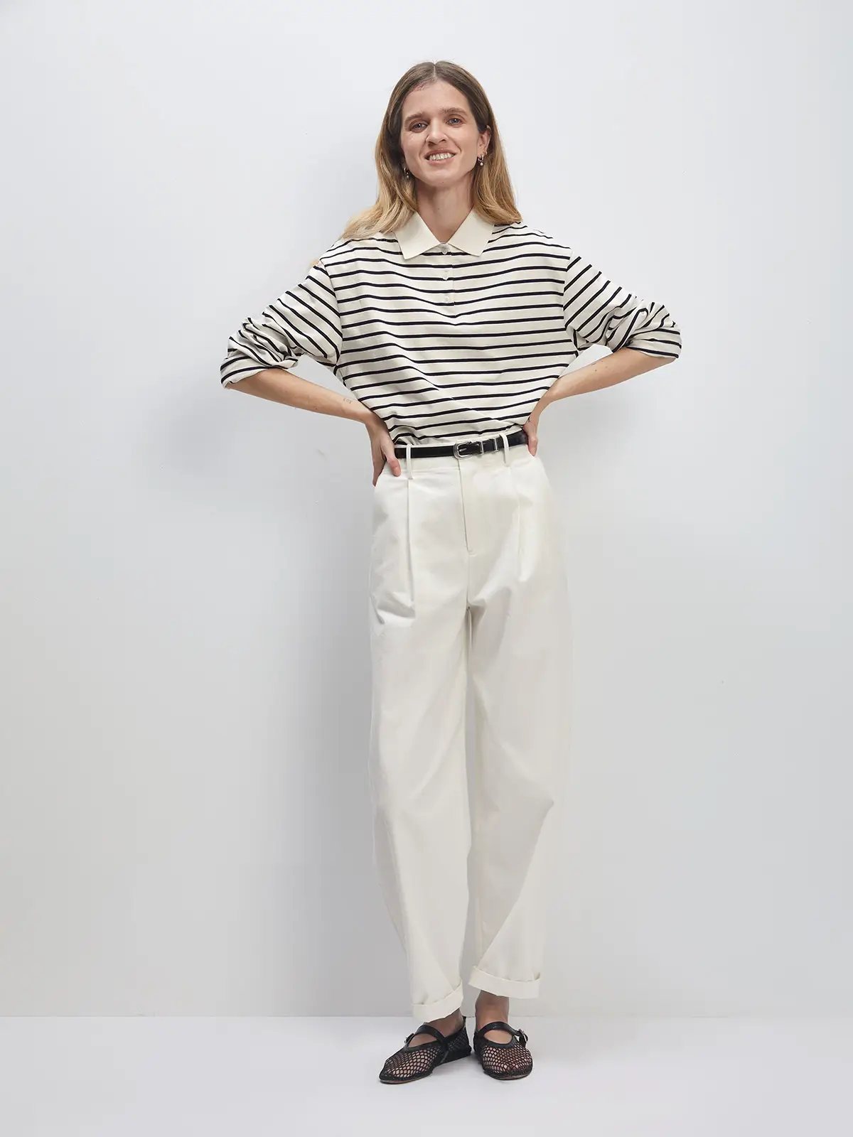 Model wearing a white/black striped long-sleeve polo collar cotton t-shirt with white trousers, hands on hips, and black mesh flats.