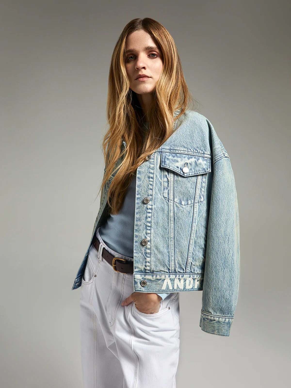 Close-up of andreatang light-blue denim jacket front showing metal buttons and logo detail at hem
