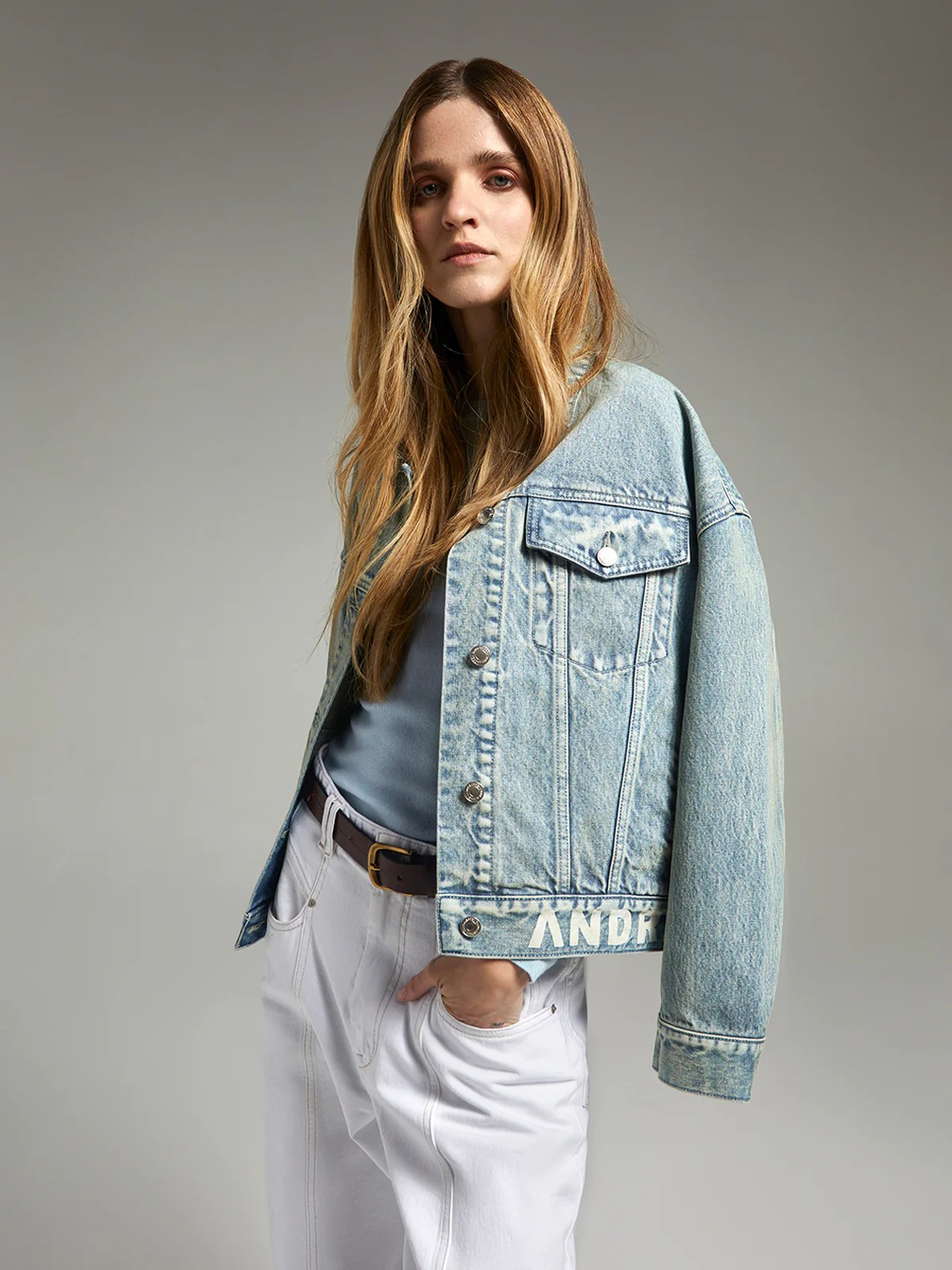 Close-up of andreatang light-blue denim jacket front showing metal buttons and logo detail at hem