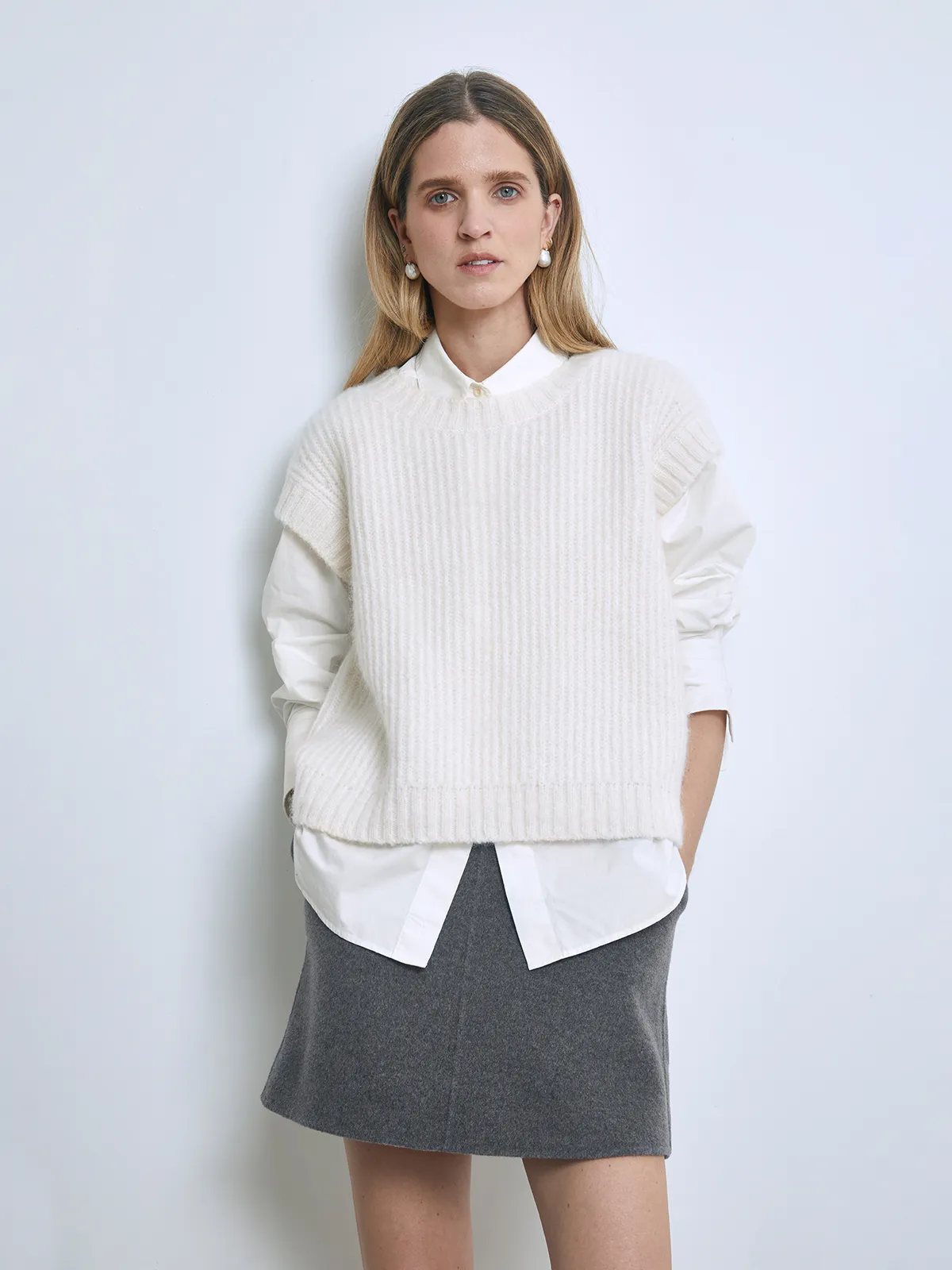 Front view of a model wearing an off-white ribbed knit vest layered over a white collared shirt and skirt.
