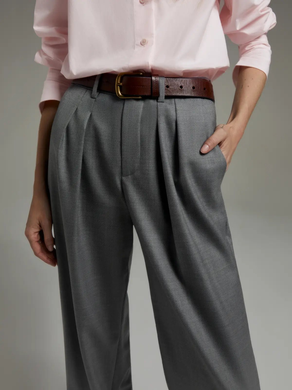 Close-up of Grey classic wool wide-leg trousers with pleats, shirt, belt, and hand in pocket.