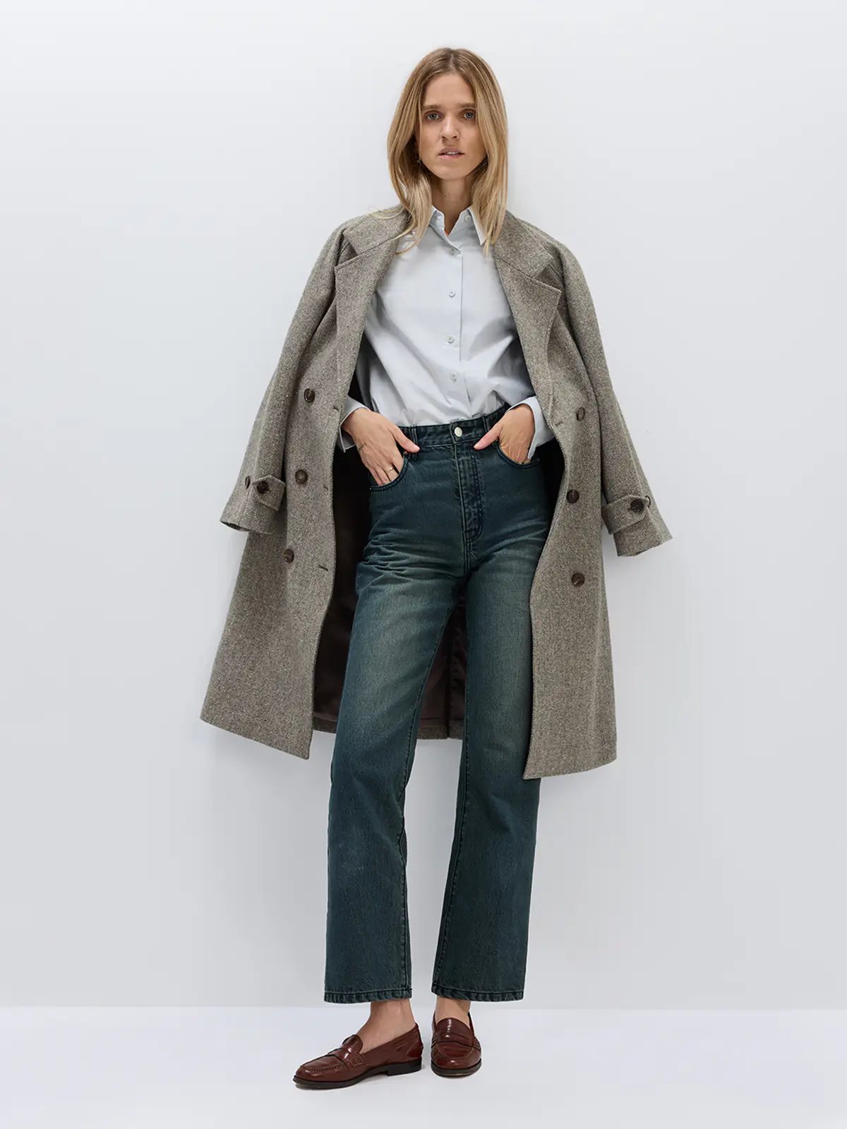 Front view of a brown herringbone double-breasted coat worn open over a sweater and trousers, holding a bag.