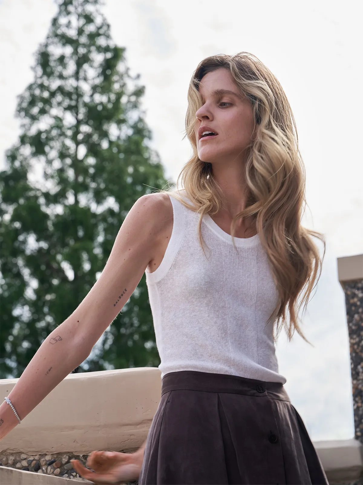 Model in ivory MEADOW Textured Knit Tank Top with brown pleated skirt, outdoor portrait showcasing lightweight drape and quiet luxury aesthetic