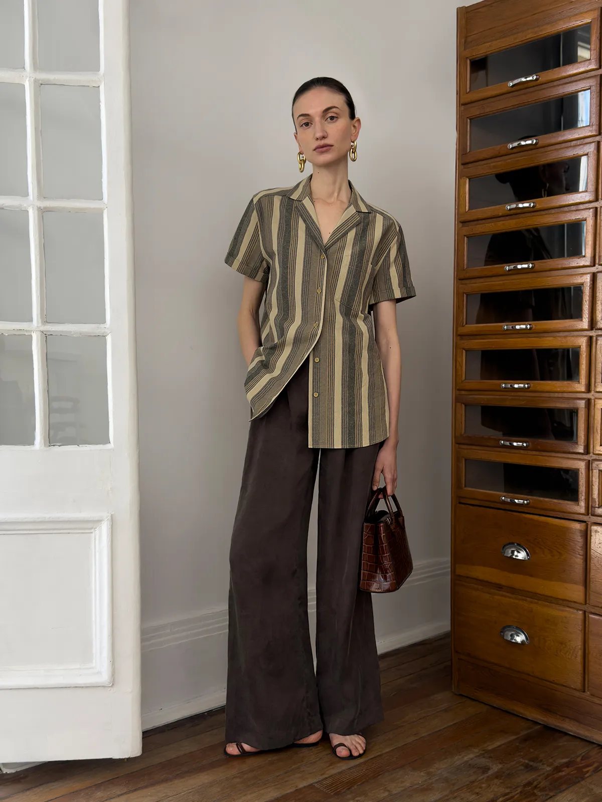 Full-length model pose in REAGAN shirt with brown trousers and croc bag beside vintage cabinet