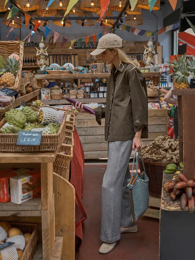 Model wearing olive green HUCK jacket in vibrant market setting, holding eggplant, styled with beige cap and woven tote