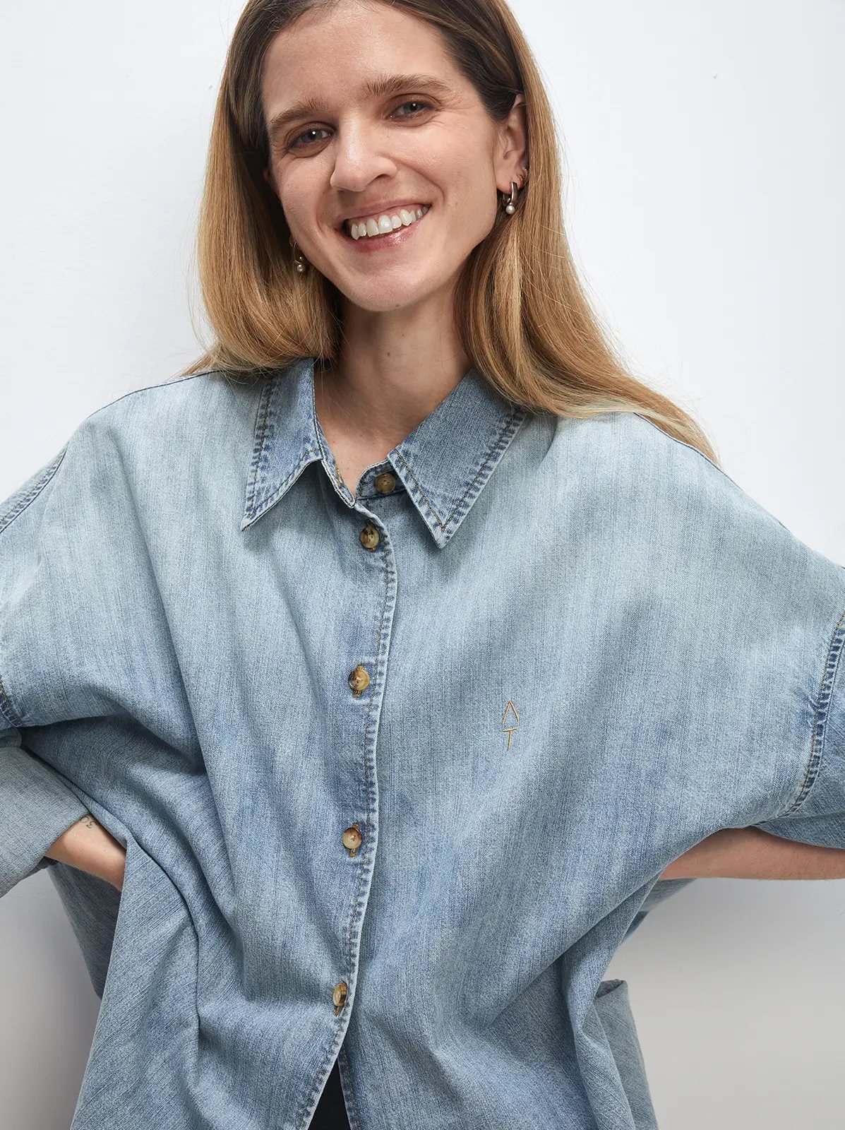 Close-up front view of a model in a light blue oversized denim shirt featuring a small embroidered logo on chest.