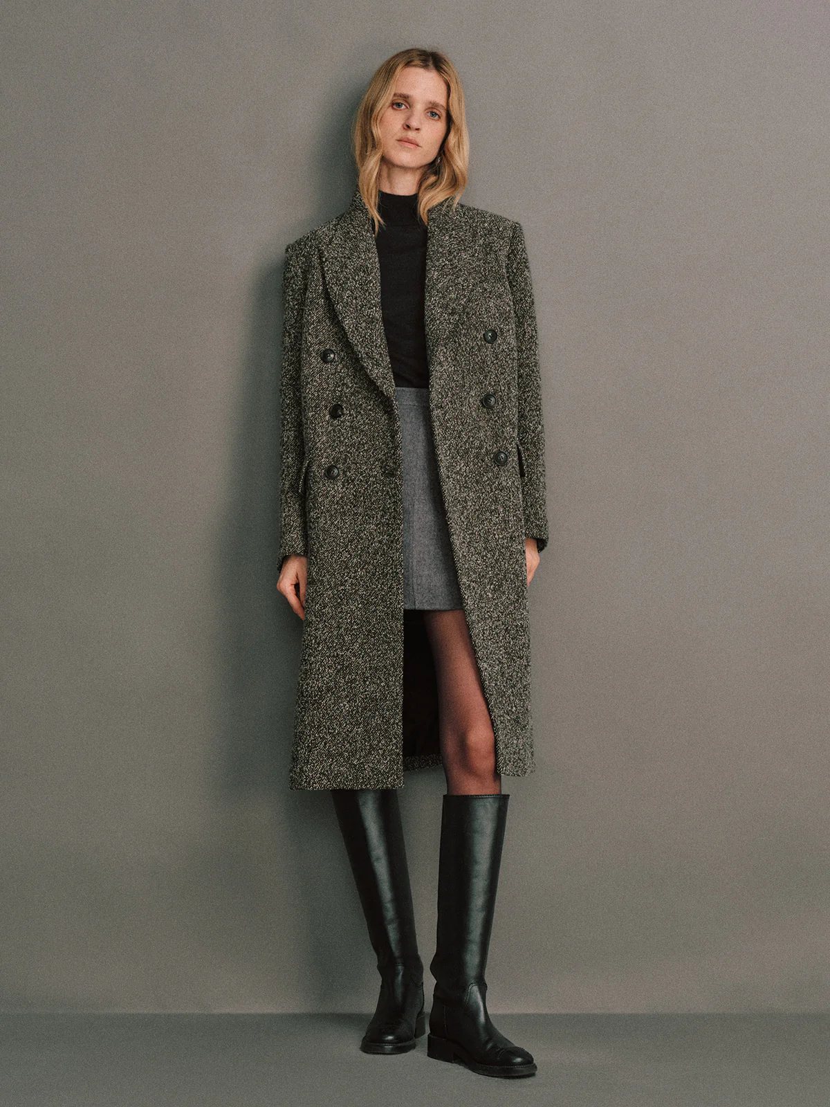 Model in a Gray wool bouclé tweed tailored long coat, double-breasted, layered over a top and denim skirt, with knee-high boots.