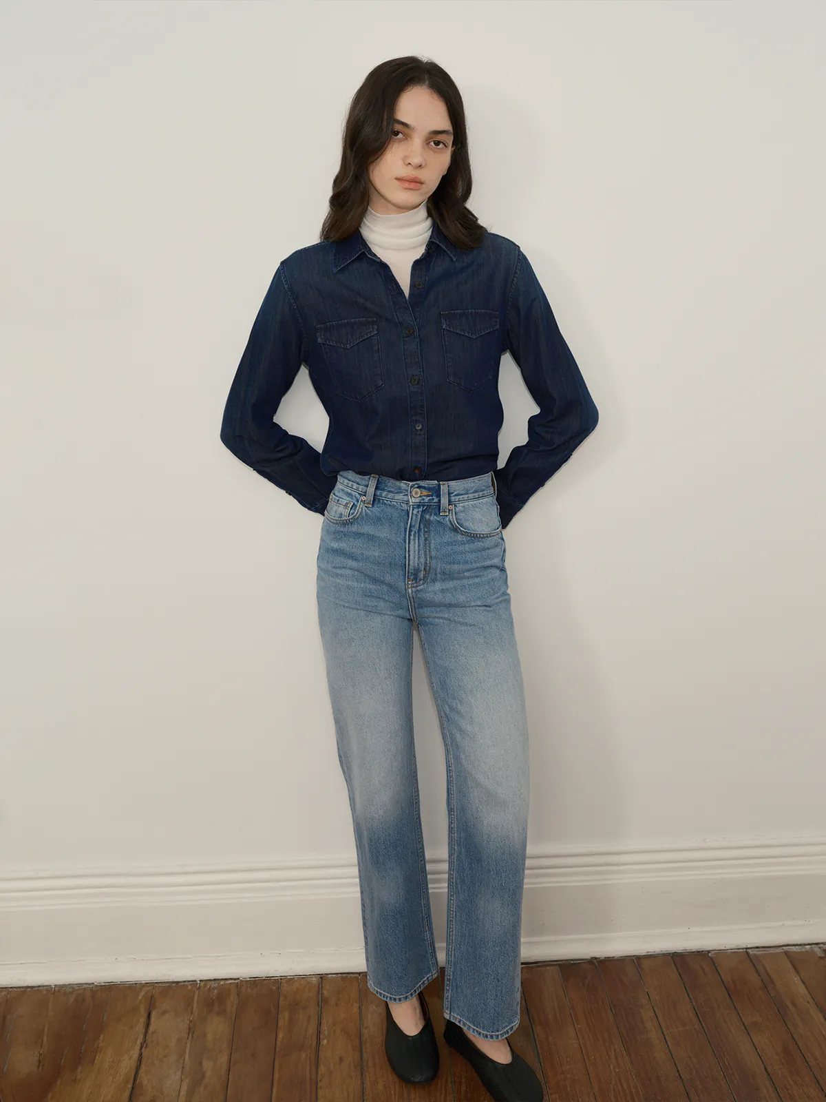 Front full-body shot of model in CINDY denim shirt with hands in pockets, light wash jeans, minimalist interior