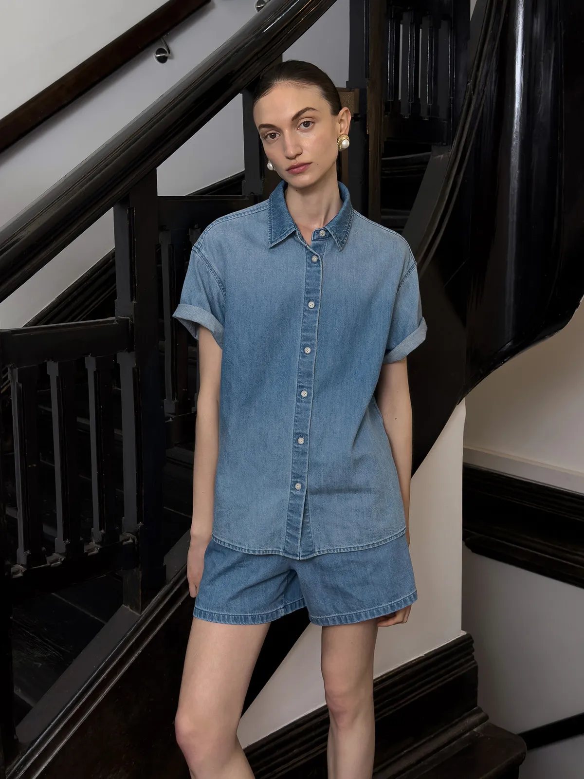 Model standing on dark wooden staircase wearing DENZEL denim shirt and matching shorts