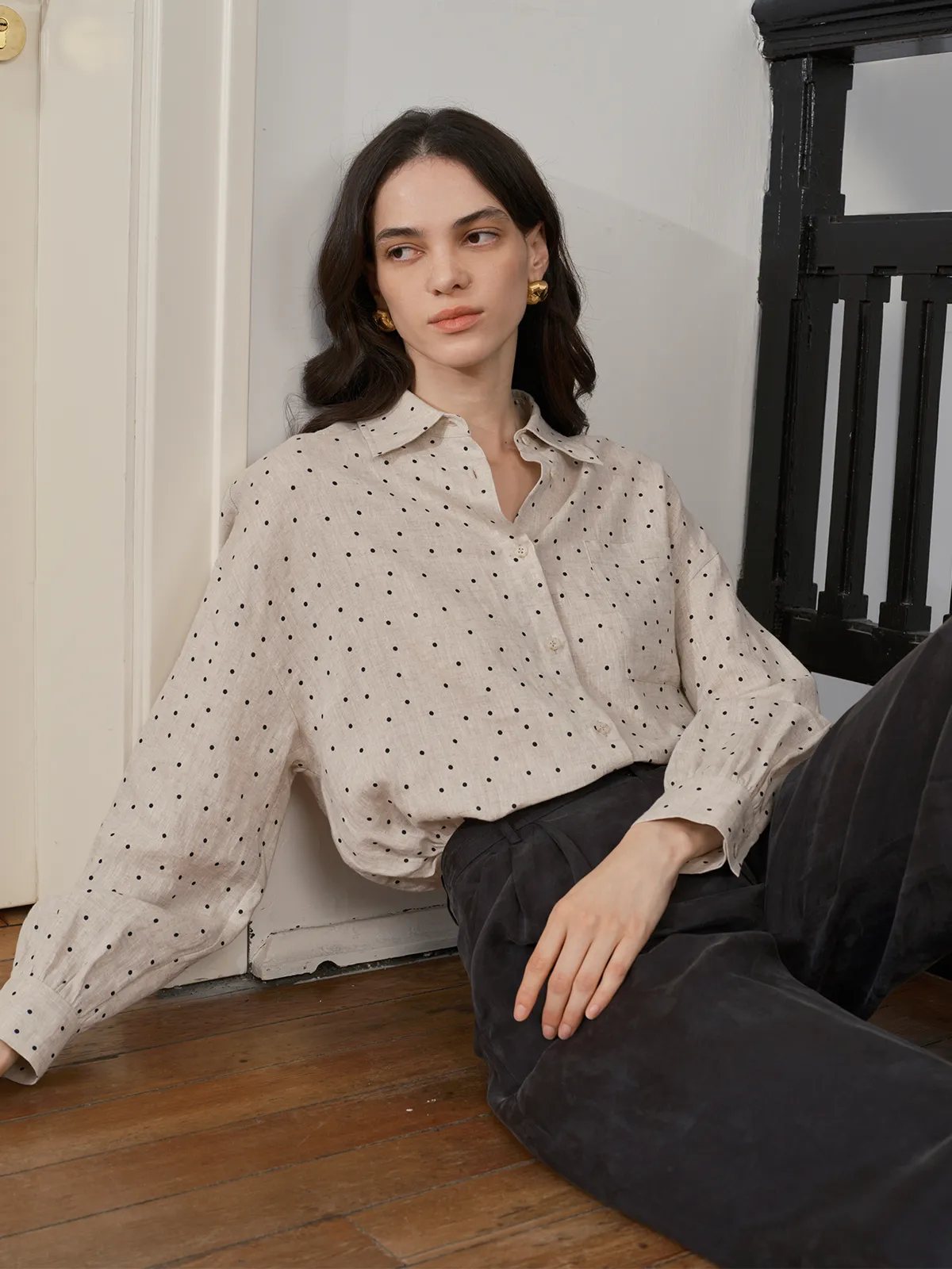 Model seated on wooden floor wearing ODELIA shirt with dark velvet trousers, highlighting sleeve drape and relaxed fit